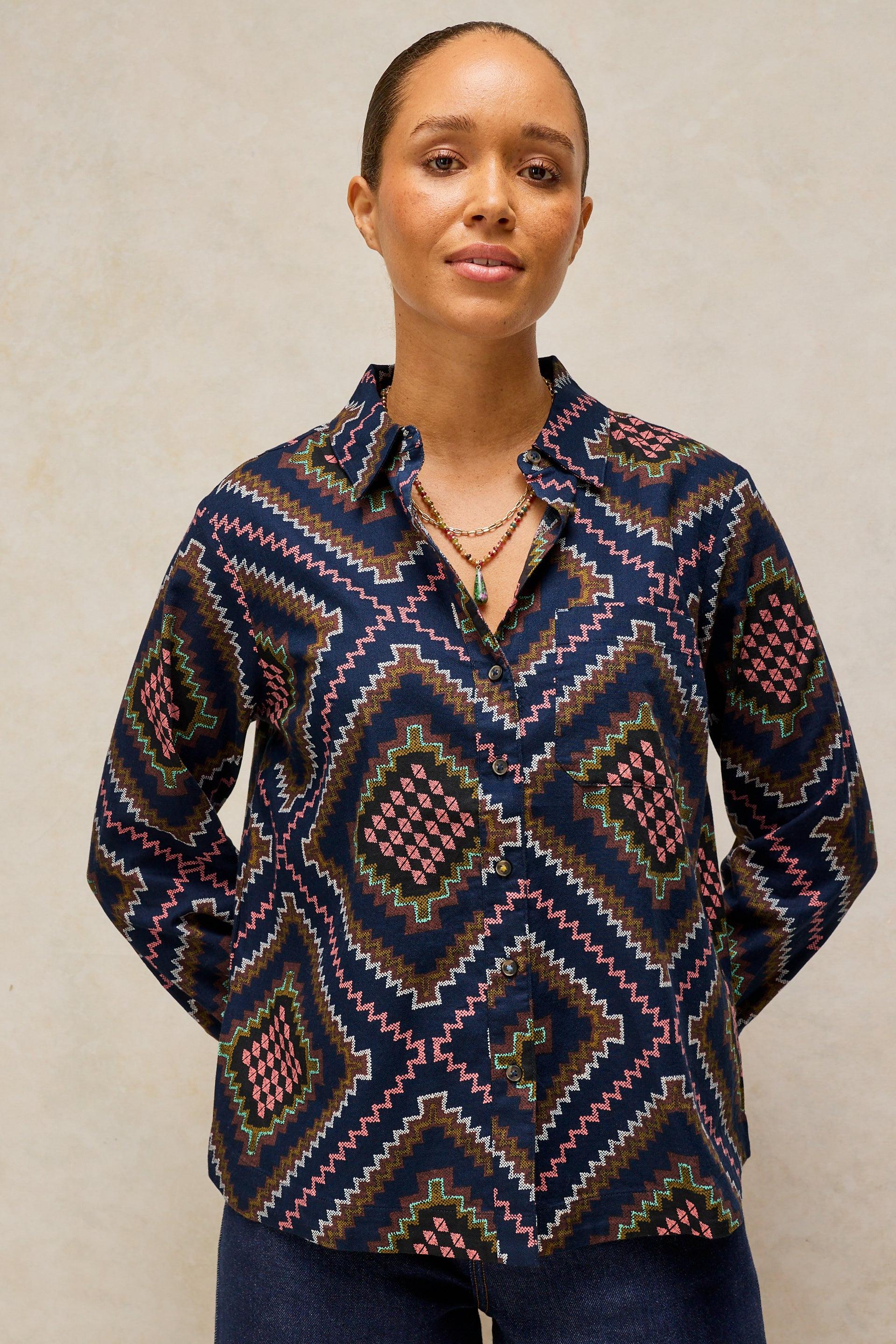 A woman wearing a long-sleeve button-up shirt with a colorful geometric diamond pattern in navy, pink, green, and brown, paired with dark blue jeans, set against a neutral textured beige background.