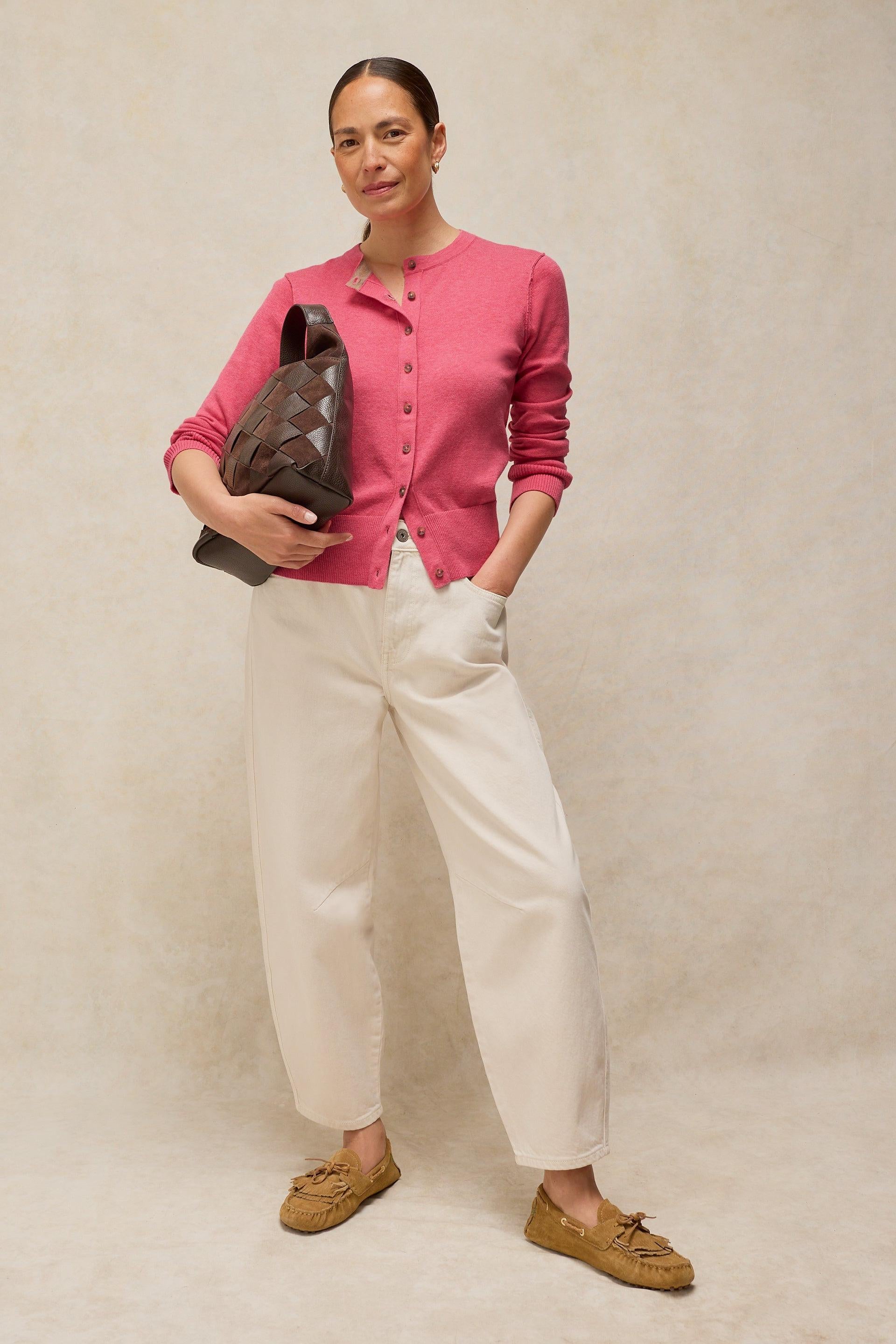 A woman posing in a pink button-up cardigan and white organic cotton trousers holding a woven brown bag