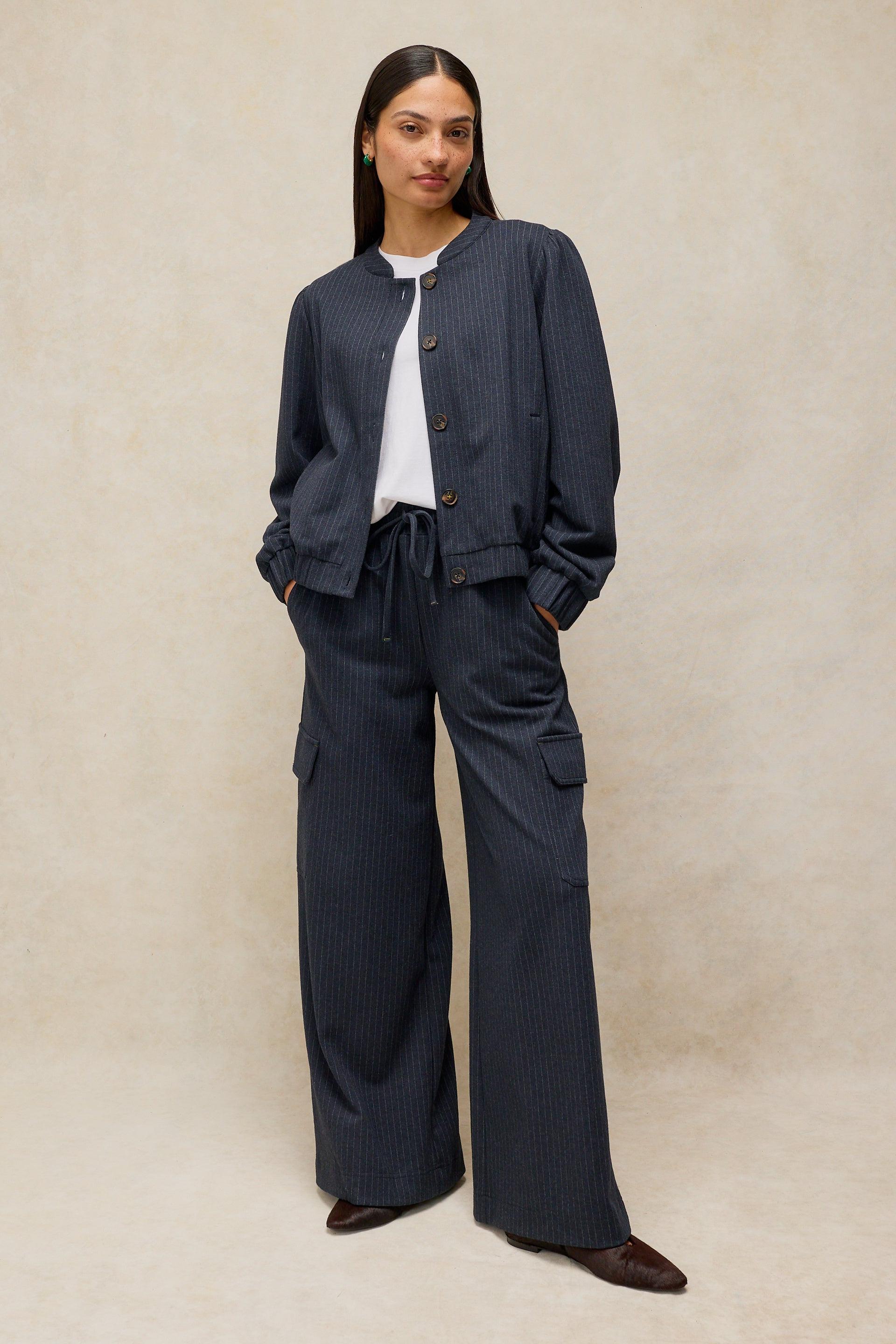 A model stands against a textured beige studio backdrop wearing a dark navy pinstriped jacket and matching wide-leg cargo pants over a white t-shirt.