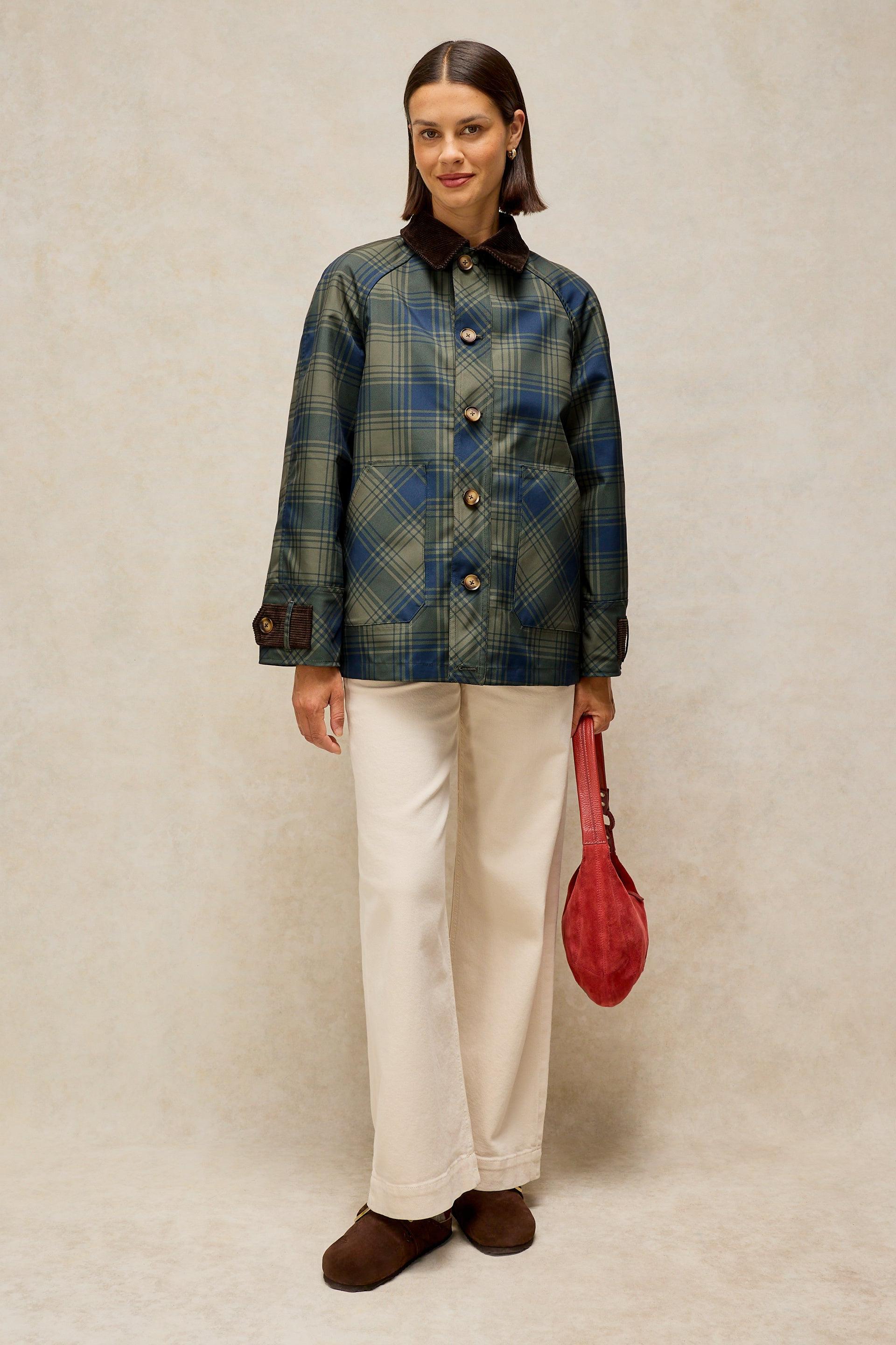A woman stands against a textured beige studio backdrop wearing a blue and green plaid button-front jacket with a dark collar, cream wide-leg pants, brown shoes, and holding a red handbag.