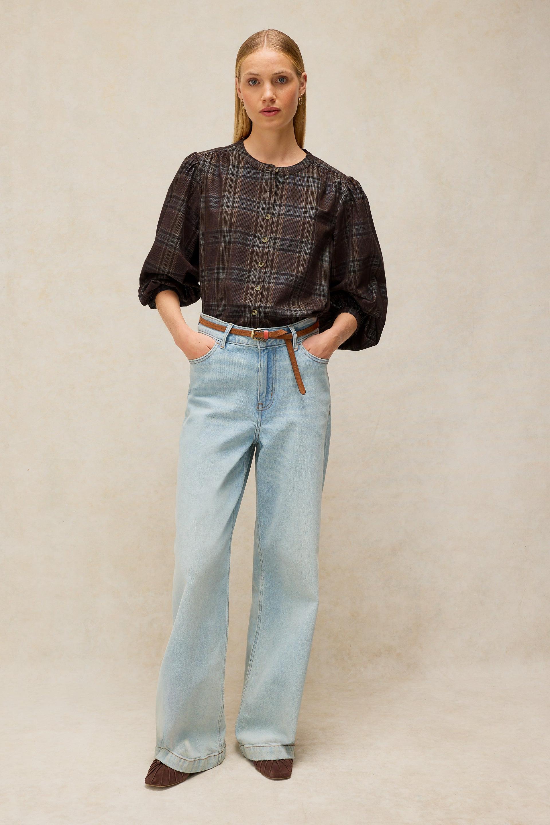 A woman stands against a textured beige studio backdrop wearing a dark brown and black plaid button-up blouse with voluminous sleeves, light-wash wide-leg jeans, and dark brown pointed shoes.