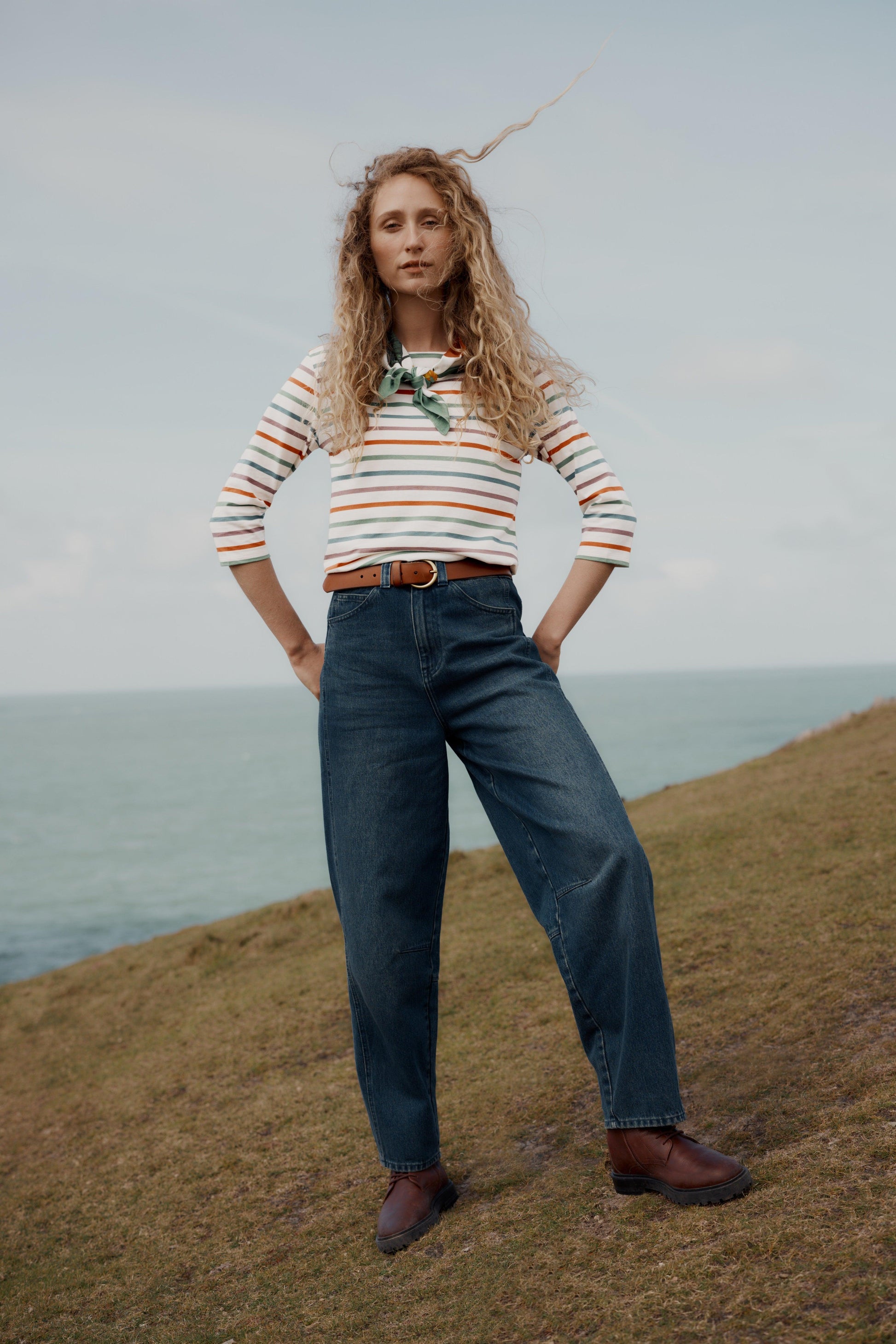 A woman stands on a grassy cliffside with the ocean and cloudy sky behind her, wearing a cream long-sleeve striped shirt with orange and green stripes, dark blue wide-leg jeans, a brown belt, and brown shoes.