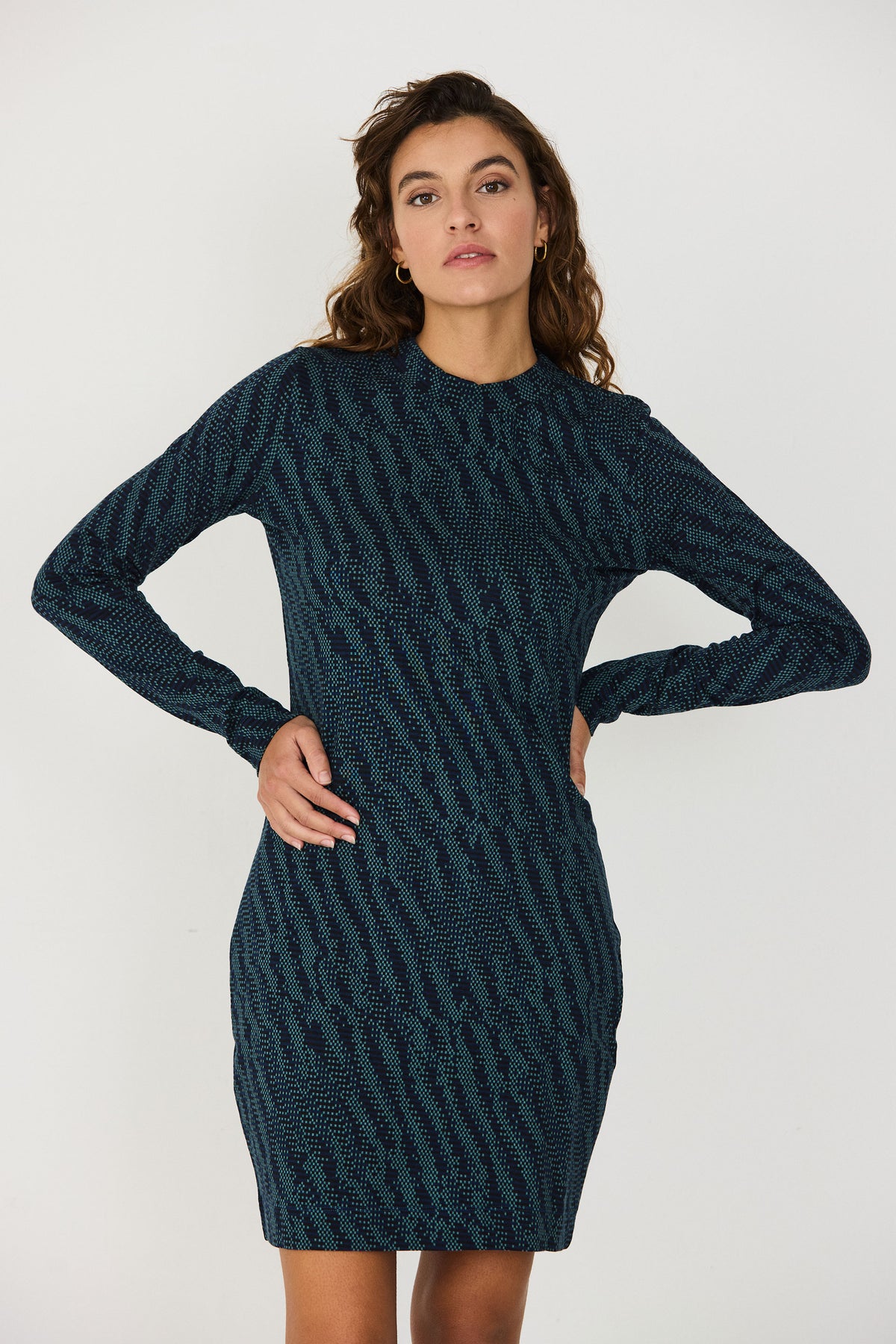 A woman posing with hands on hips wearing a fitted, patterned long-sleeve green dress against a white background.