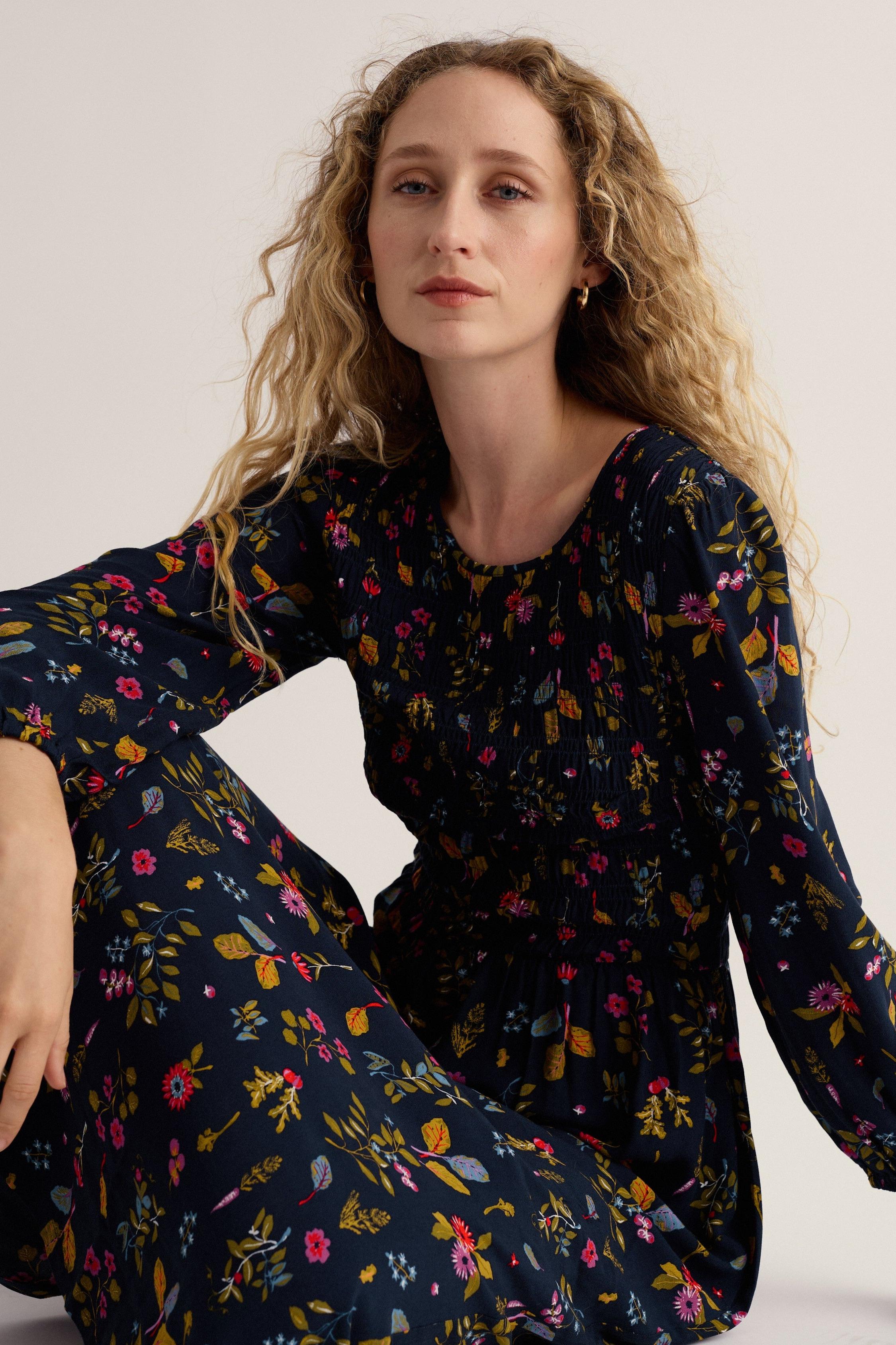 A woman with curly blonde hair wearing a dark navy floral jumpsuit, sitting against a plain light beige studio background.