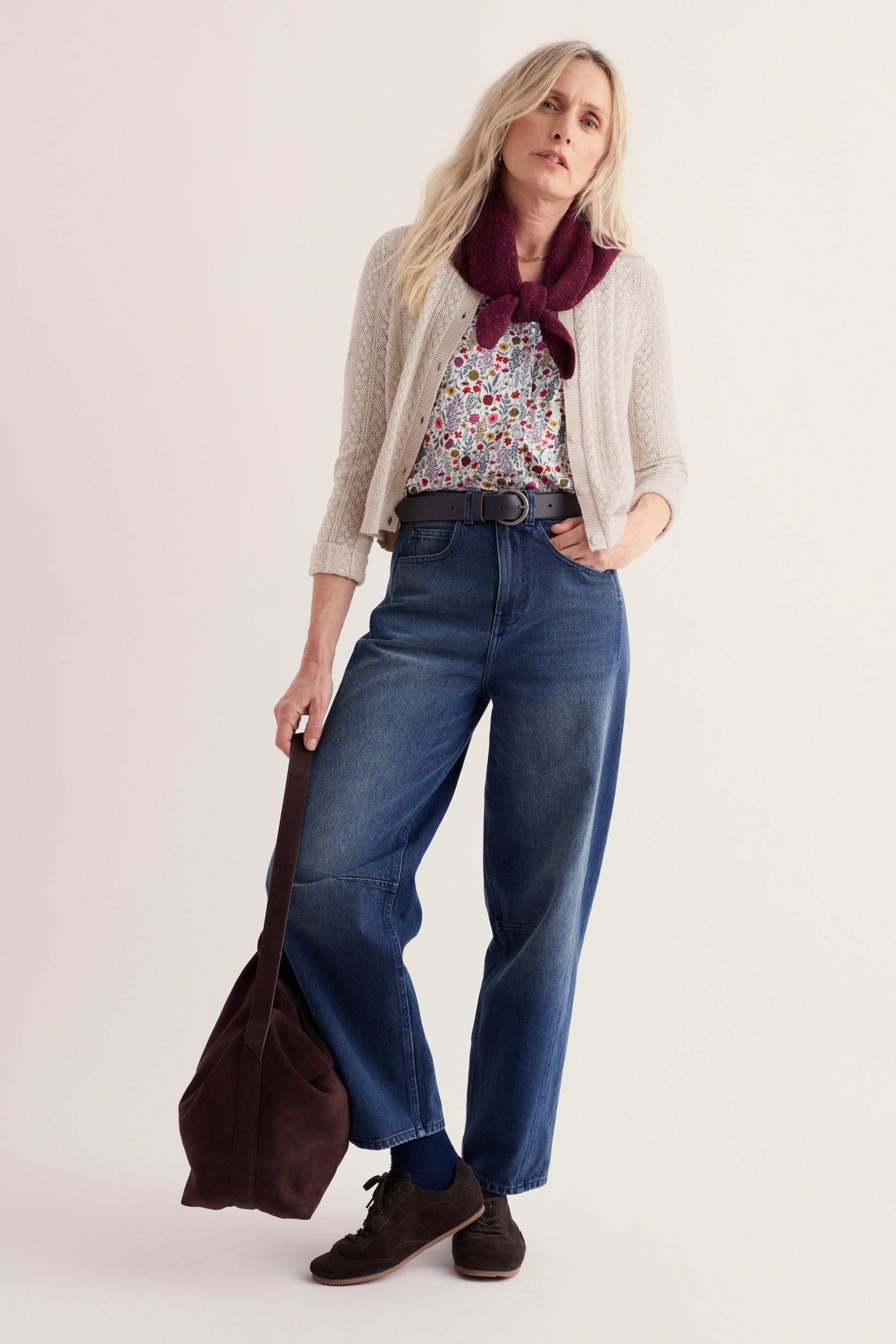 A woman modeling a light beige knit cardigan, a floral print top, a dark burgundy scarf, blue wide-leg jeans, and dark brown shoes, holding a dark brown tote bag against a plain light beige studio background.
