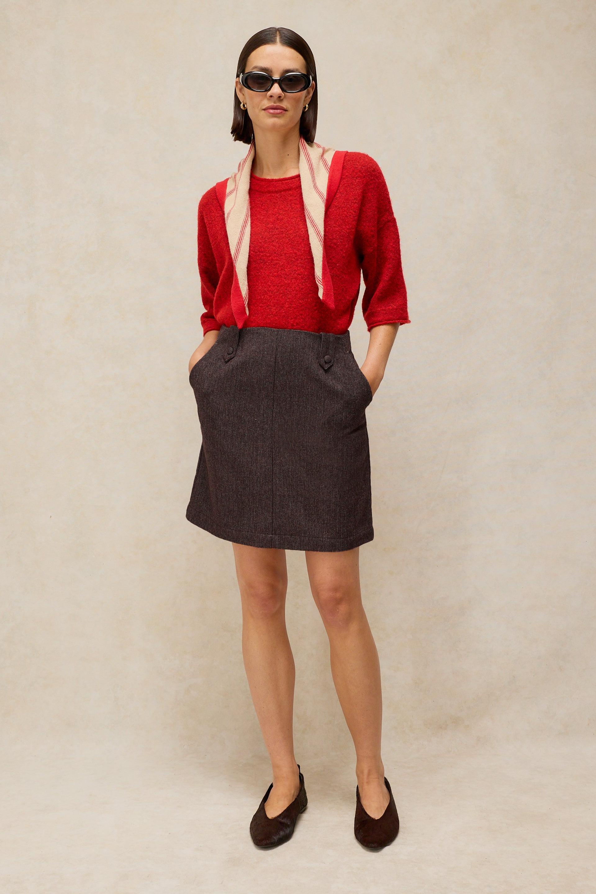 A model stands against a textured beige backdrop wearing a red knit top, a plum textured mini skirt with front pockets, a light pink scarf, and dark brown flats.