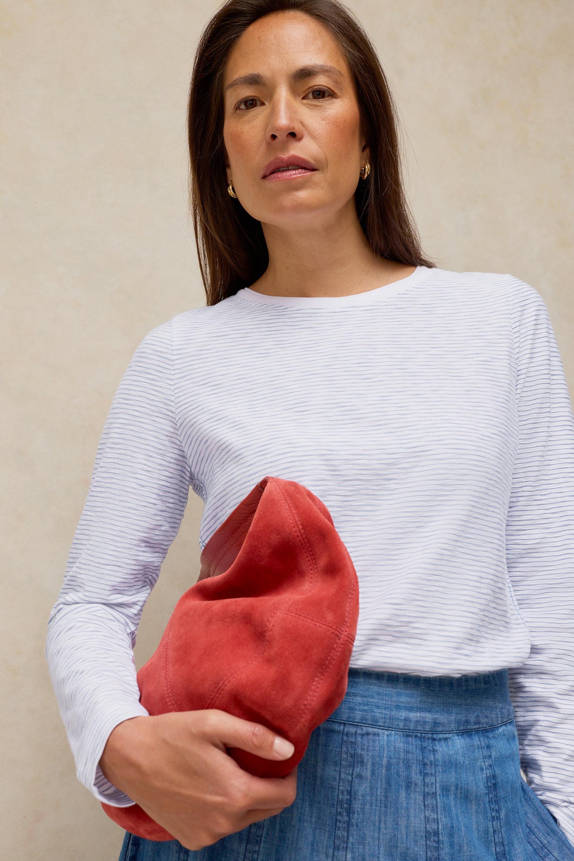 Woman wearing a striped cotton long sleeve top holding a red suede clutch bag.