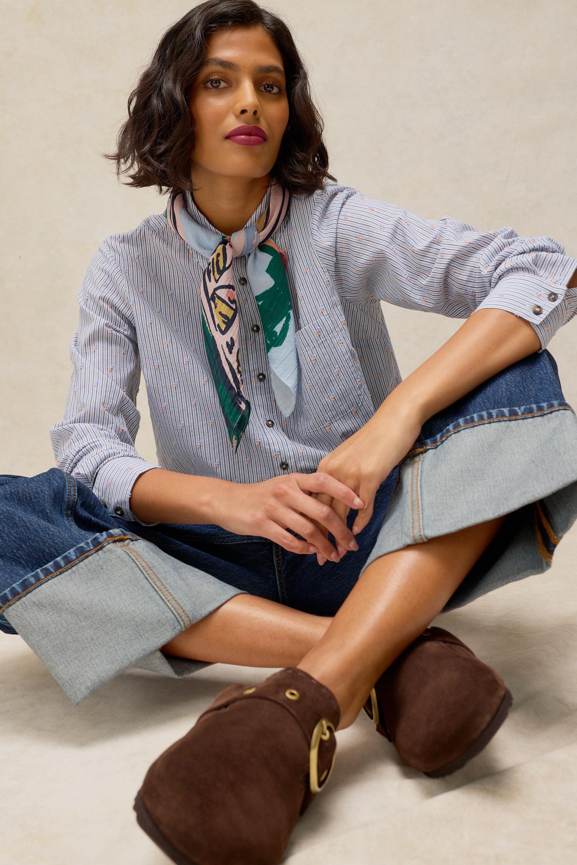 A woman sits cross-legged wearing a light blue and white striped button-up shirt with a green and white patterned silk neck scarf, blue denim shorts with rolled cuffs, and brown suede clogs with gold-tone hardware, against a neutral beige background.