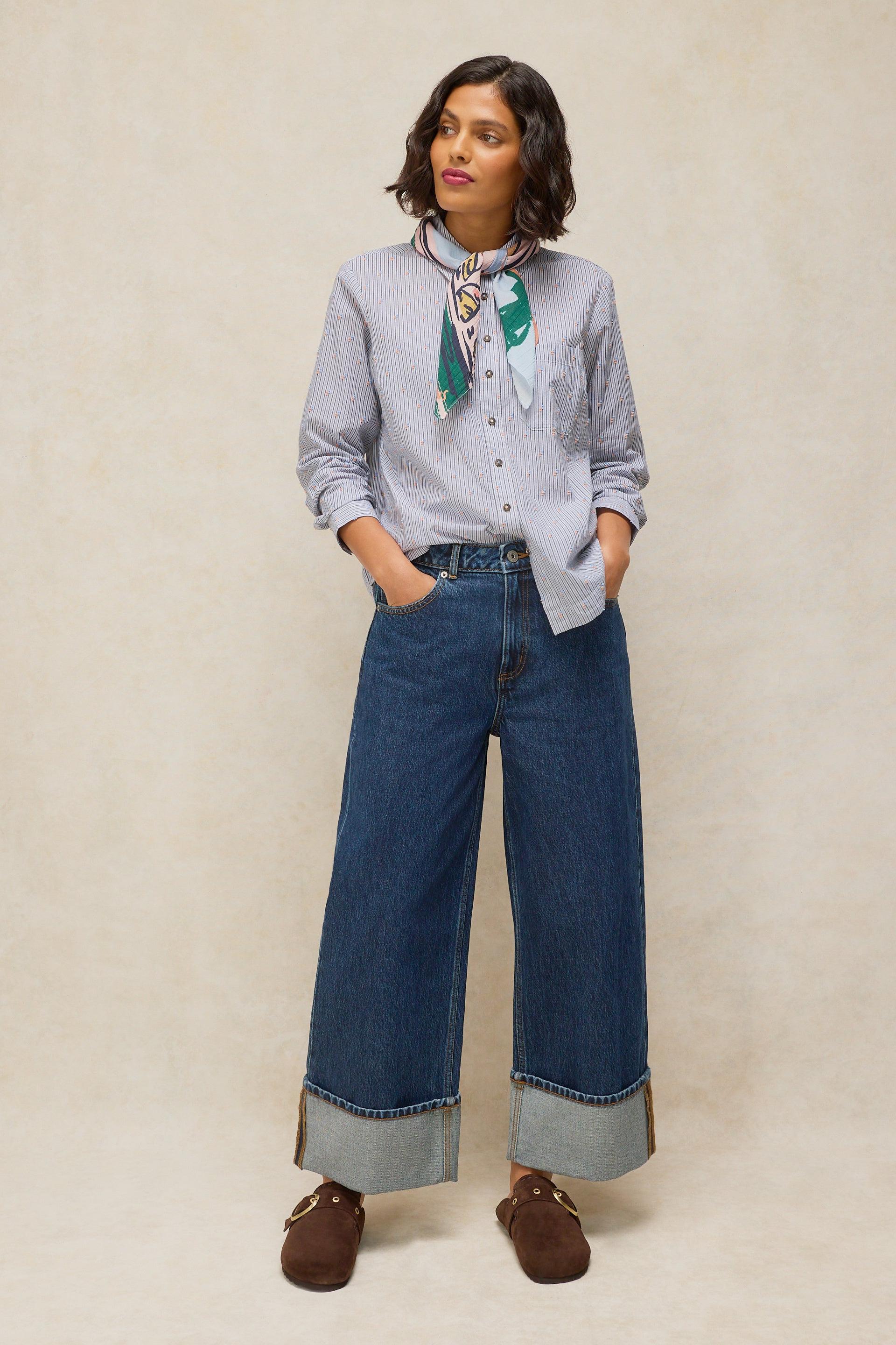 A woman modeling a light blue pinstriped button-down shirt, wide-leg blue jeans with rolled cuffs, a green and white patterned neck scarf, and brown leather loafers, set against a neutral beige textured studio backdrop.