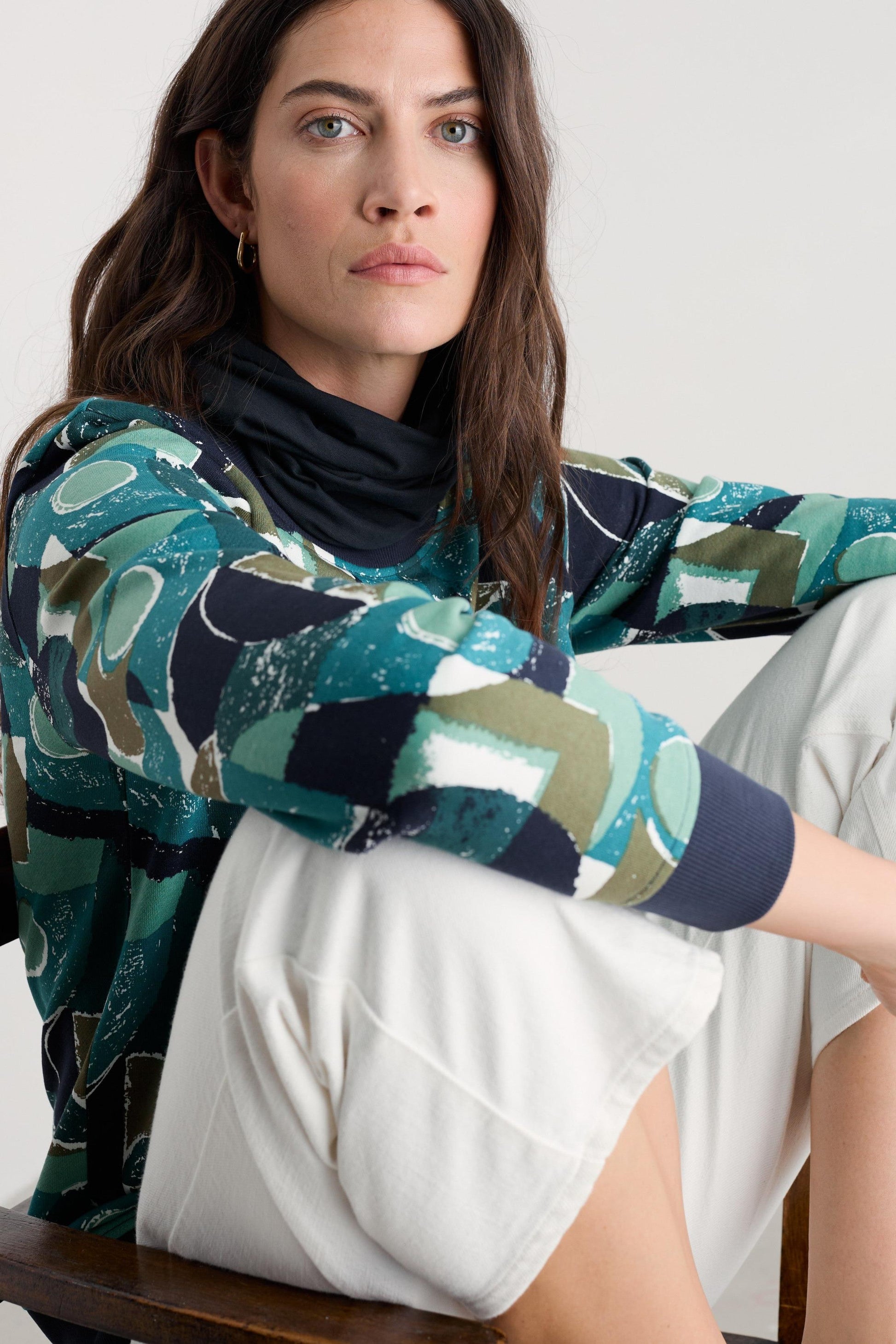 A woman seated on a wooden chair wearing a long-sleeve abstract print top with teal, navy, olive, and cream shapes over a black turtleneck, paired with white pants.