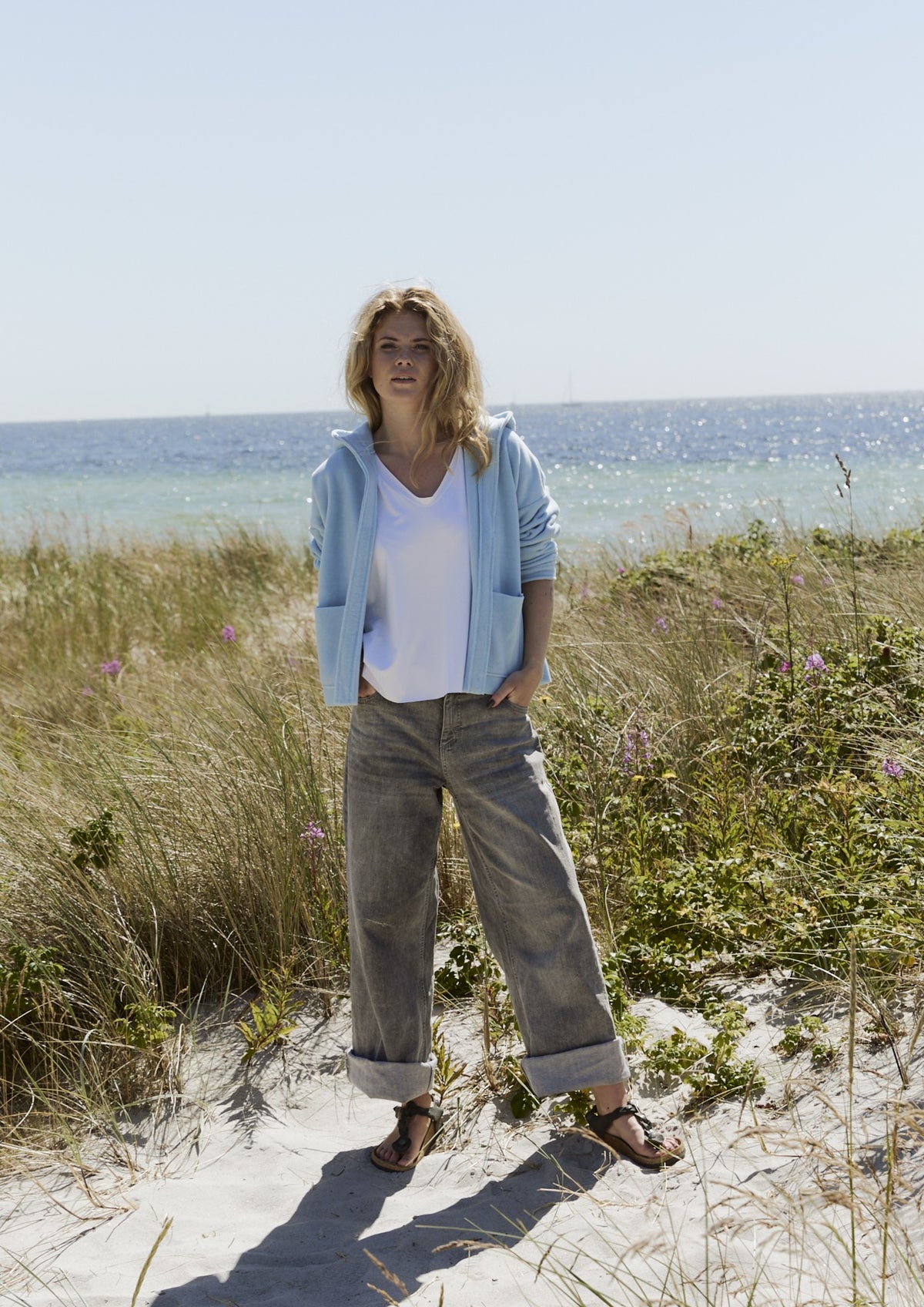 A woman stands on a sandy beach wearing a light baby blue Henriette Steffensen fleece cardigan.