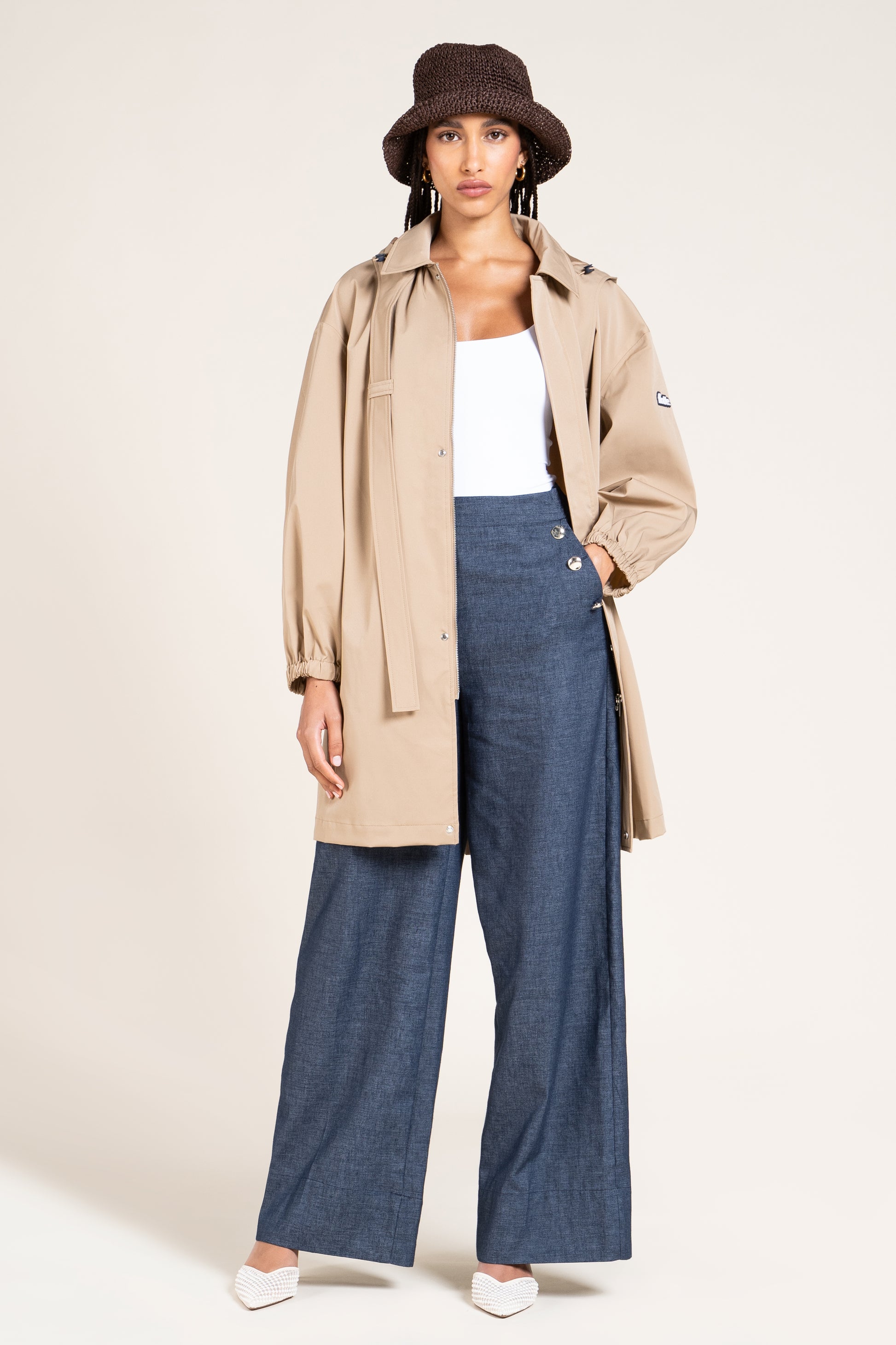 A woman wearing a beige sustainable Flotte waterproof coat and dark blue wide-leg trousers against a plain background.