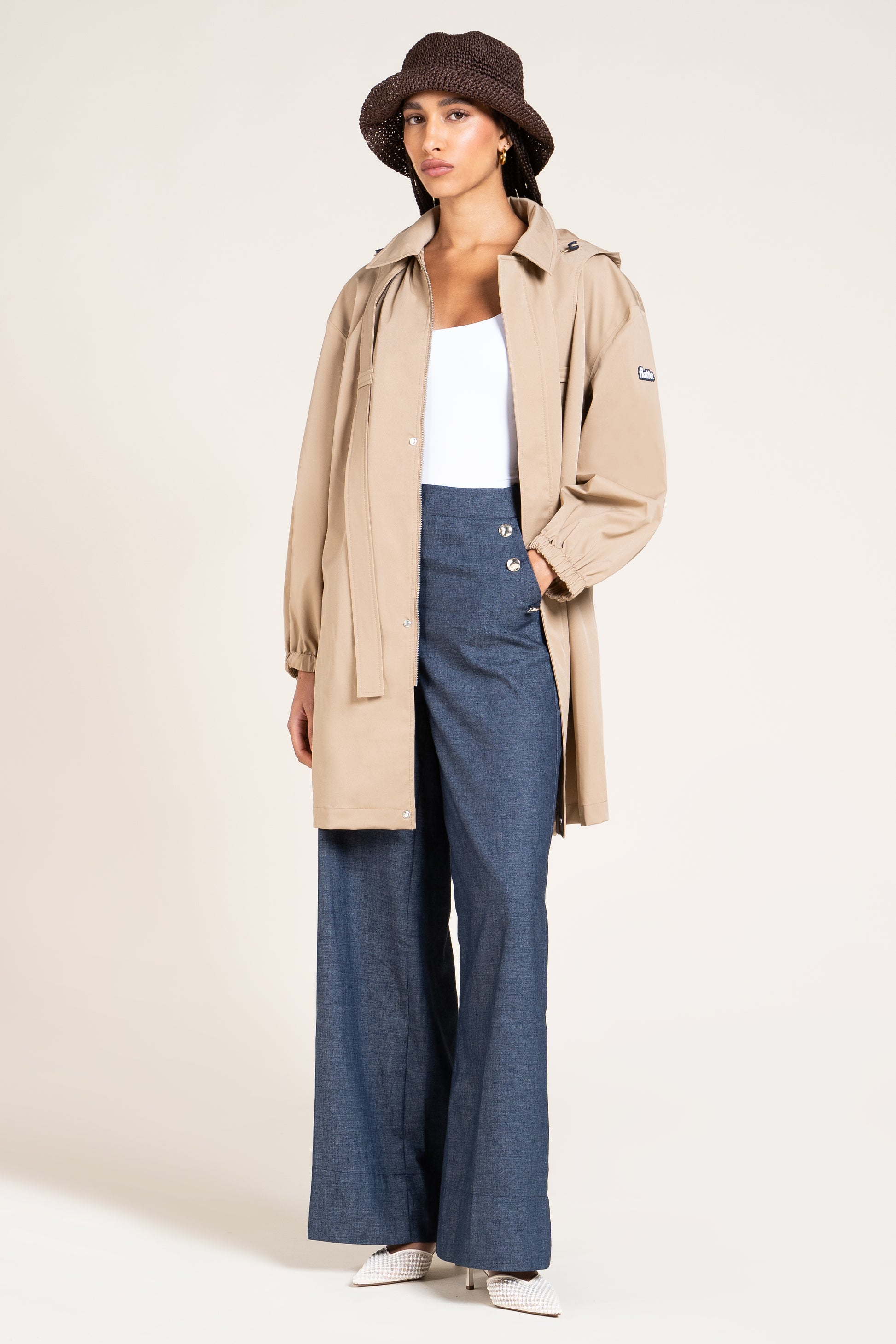 A model wearing a beige hooded trench coat over a white tank top and blue wide-leg trousers, styled with a dark brown bucket hat and white sneakers, against a plain light beige studio background.