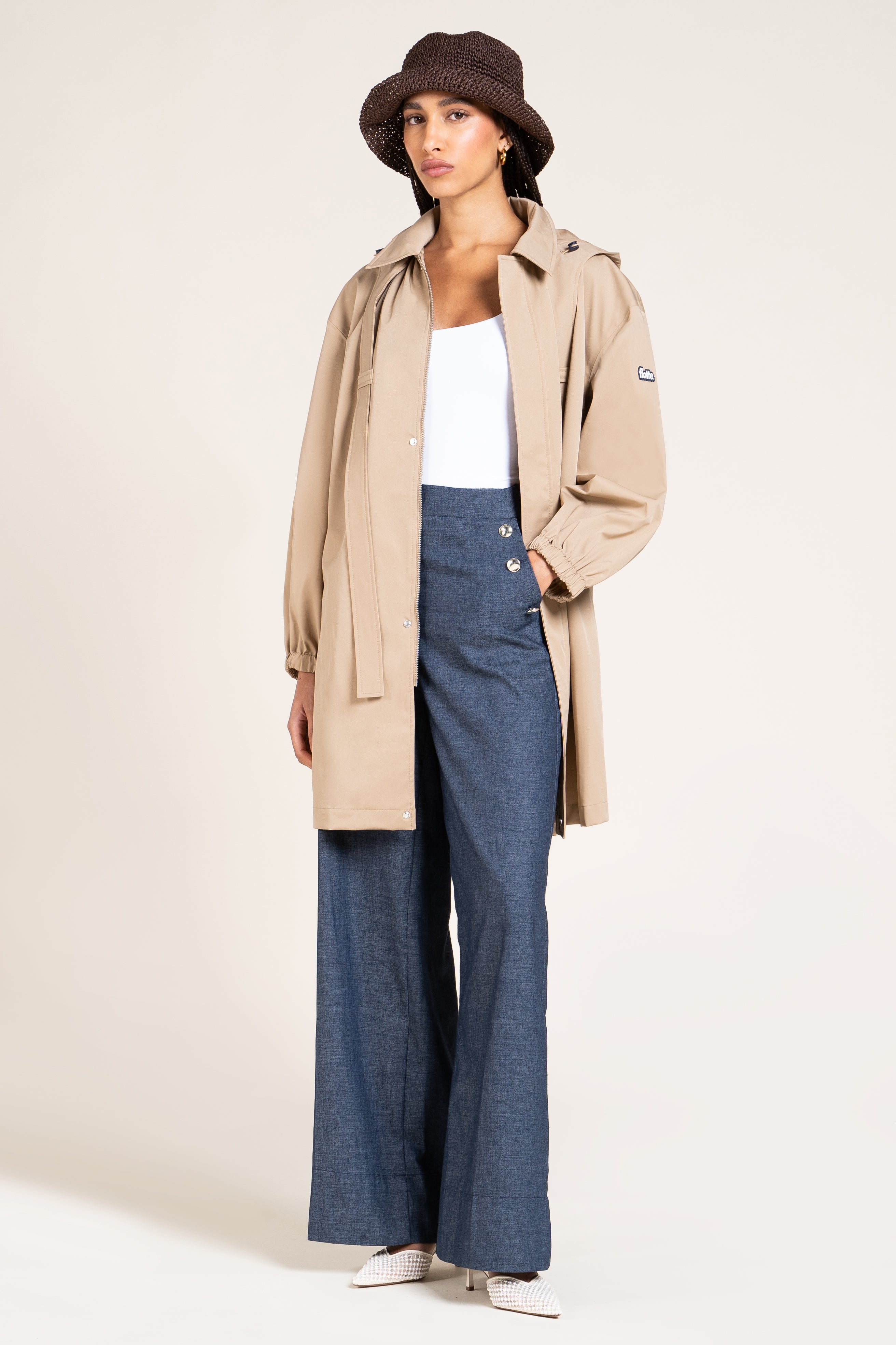 A model wearing a beige hooded trench coat over a white tank top and blue wide-leg trousers, styled with a dark brown bucket hat and white sneakers, against a plain light beige studio background.