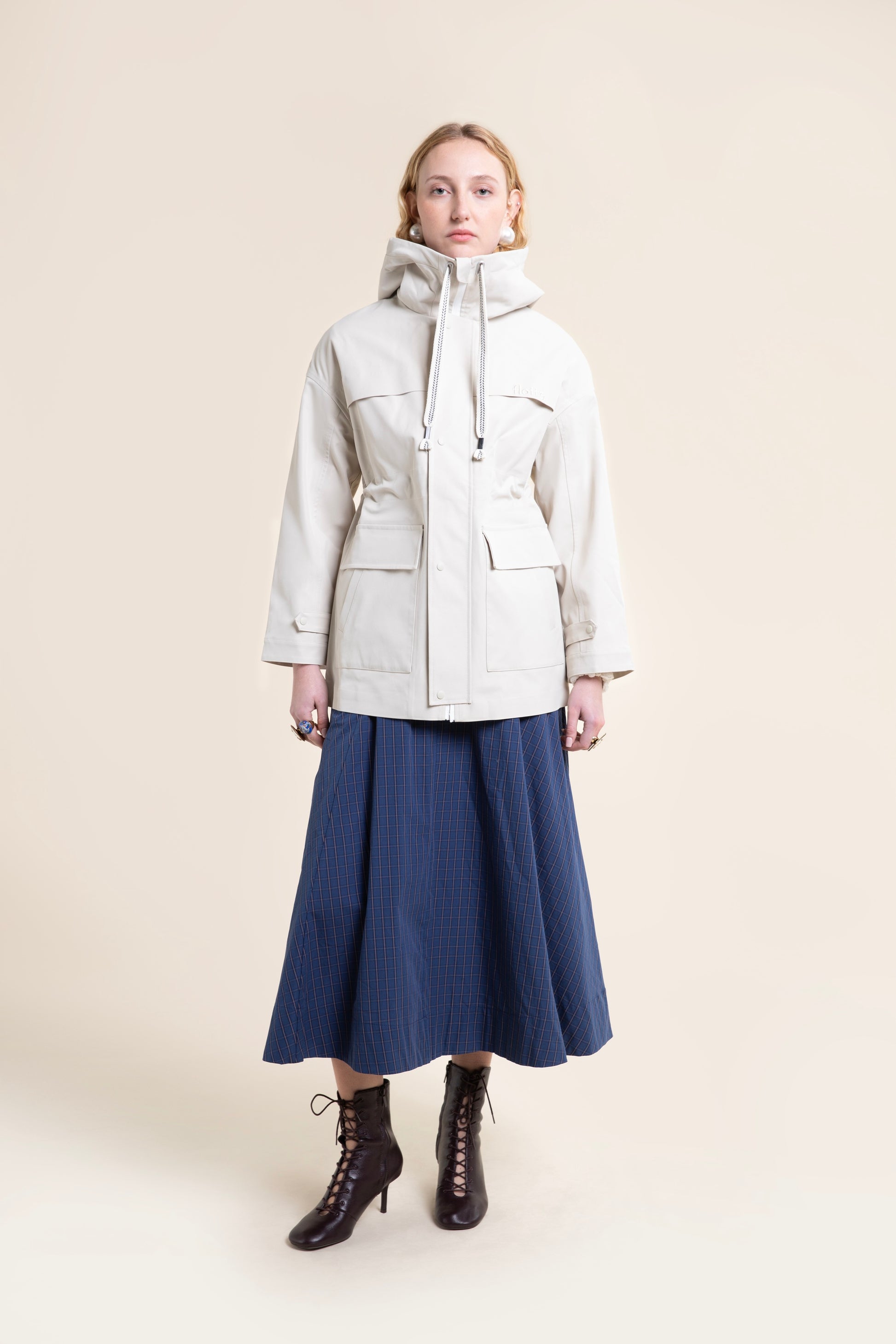 A woman modeling a beige Flotte waterproof parka coat with a blue plaid skirt and lace-up boots.