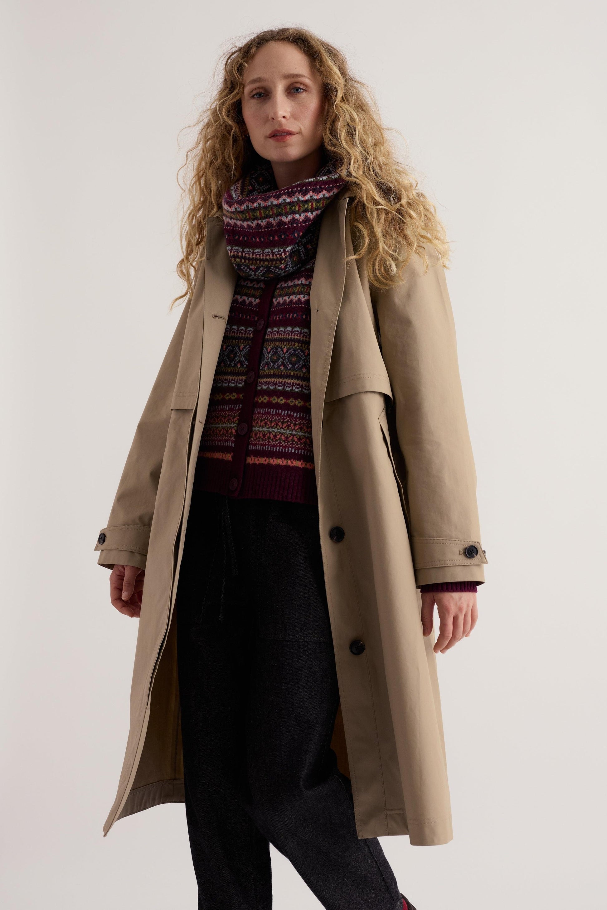 Woman wearing a beige Seasalt Cornwall trench coat layered over a patterned sweater and dark trousers.