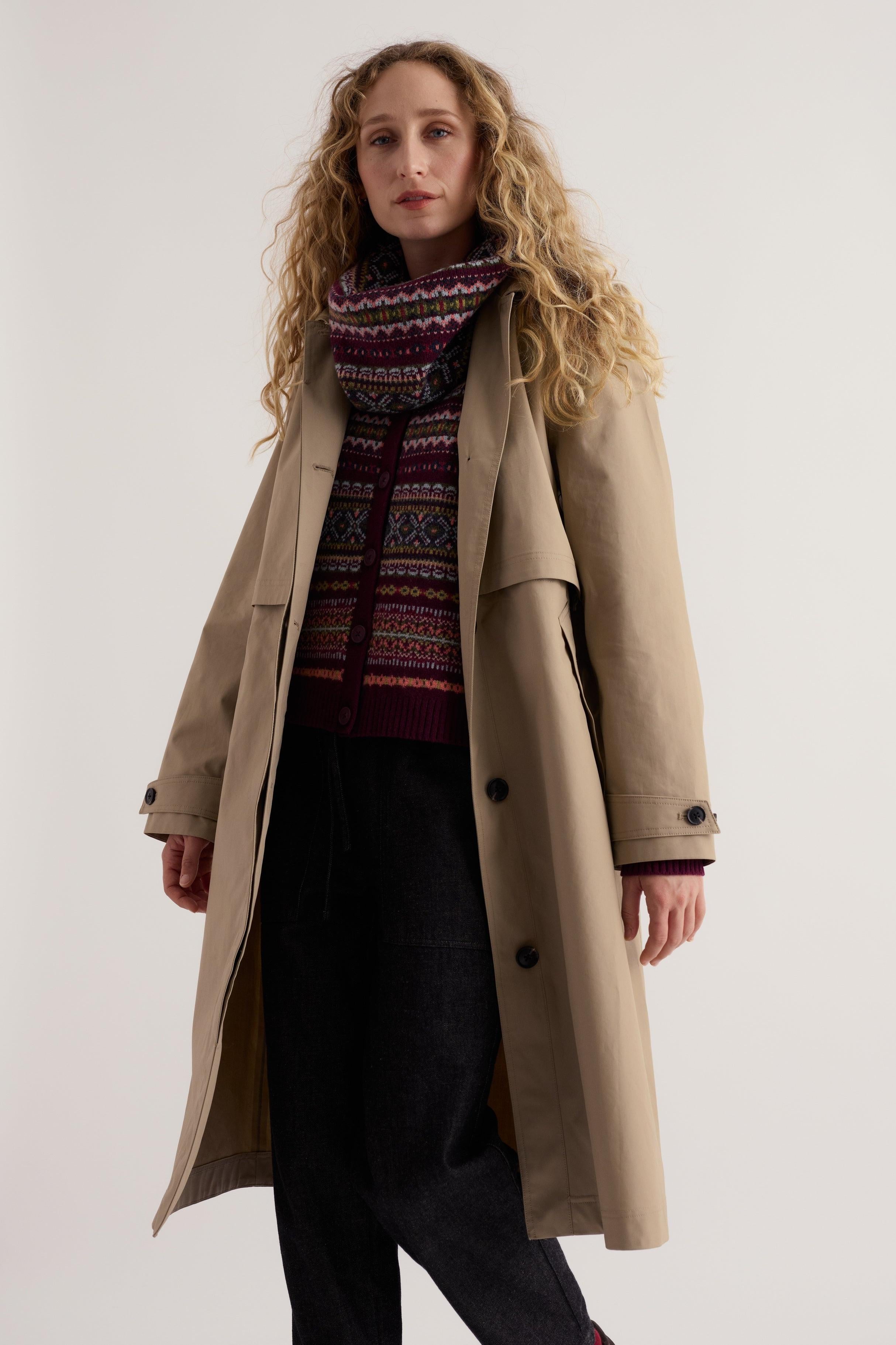 Woman wearing a beige Seasalt Cornwall trench coat layered over a patterned sweater and dark trousers.