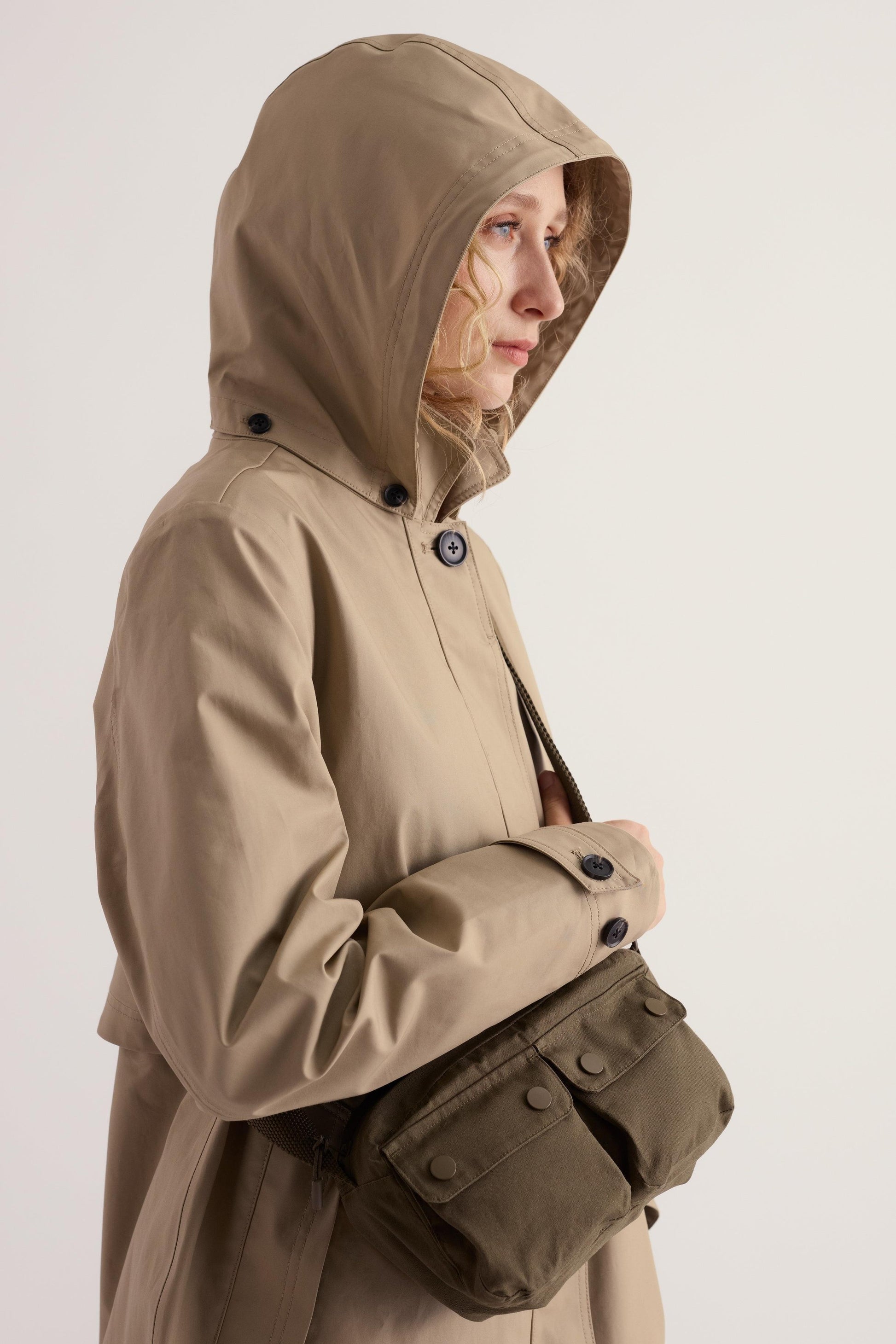 Woman wearing a beige waterproof Seasalt Cornwall trench coat with a hood up and an olive green crossbody bag.
