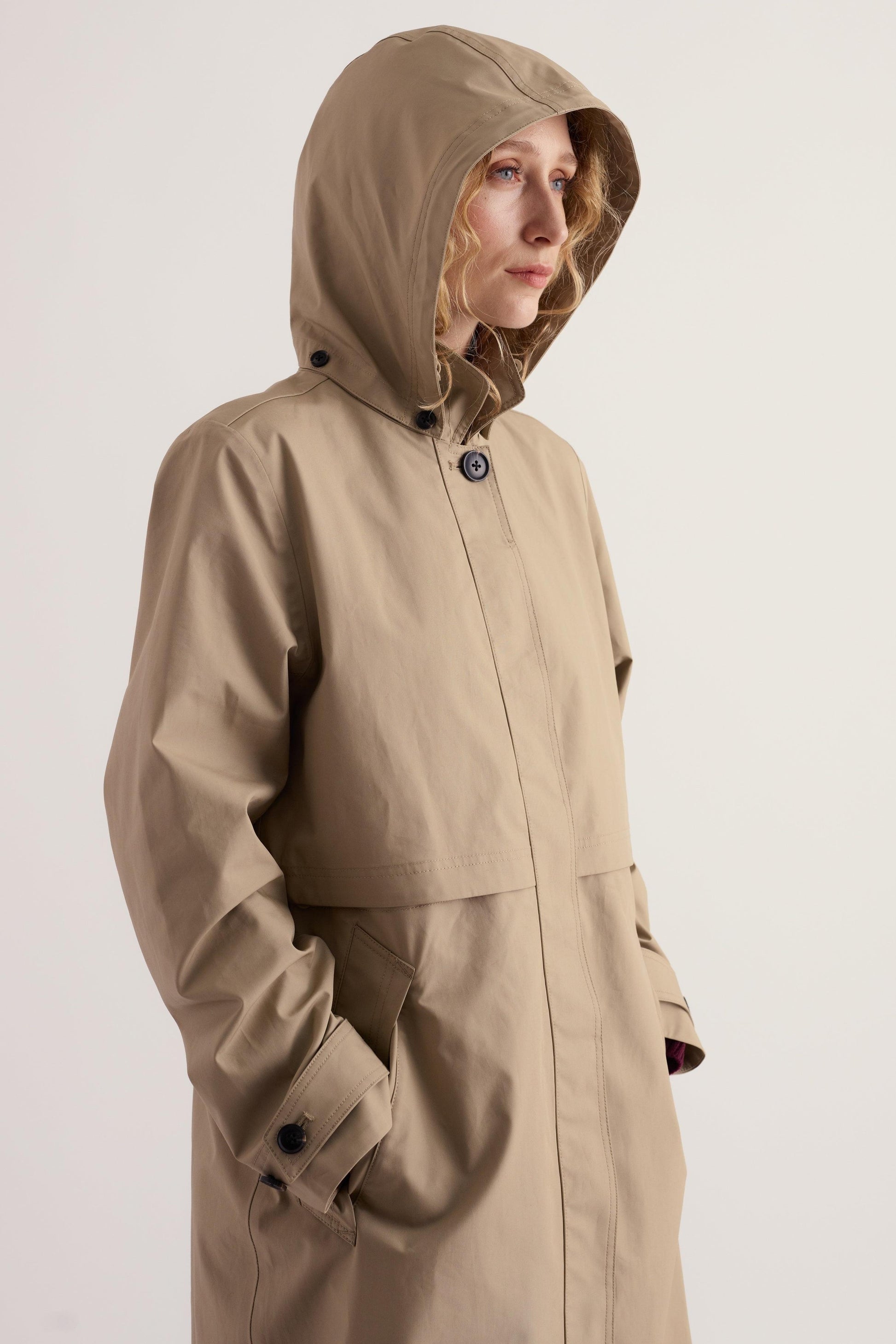 A woman wearing a beige hooded Seasalt waterproof trench coat with a clean, light-colored background.