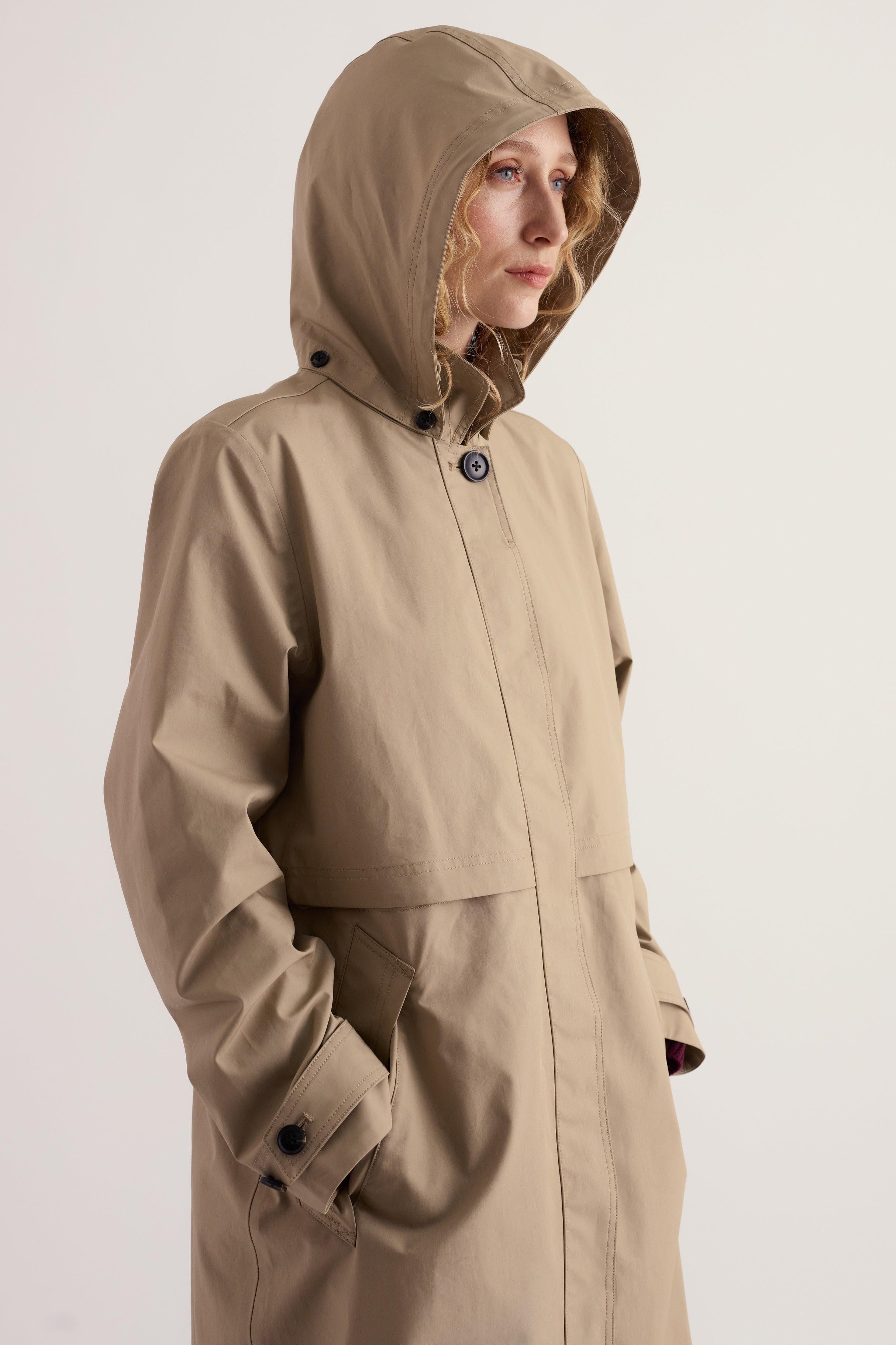 A woman wearing a beige hooded Seasalt waterproof trench coat with a clean, light-colored background.