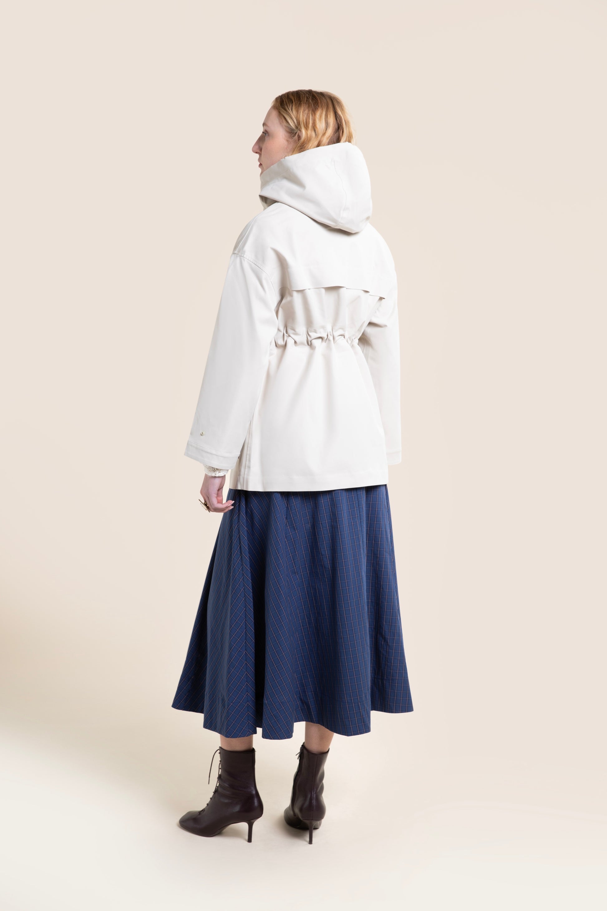 A woman wearing a stylish beige hooded waterproof coat over a long blue plaid skirt against a neutral background.