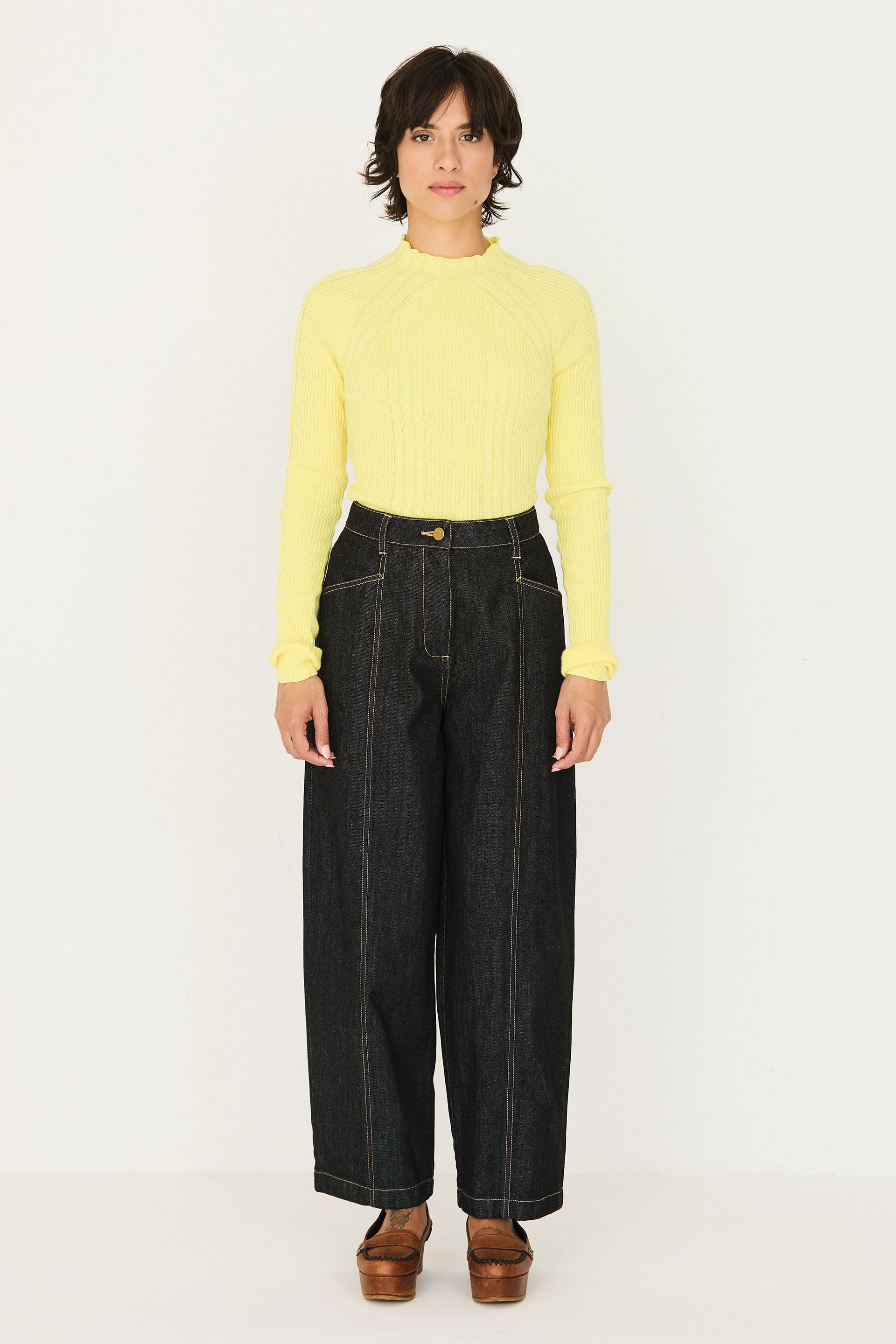 A woman wearing a bright yellow ribbed sweater and black barrel leg jeans standing against a white background.