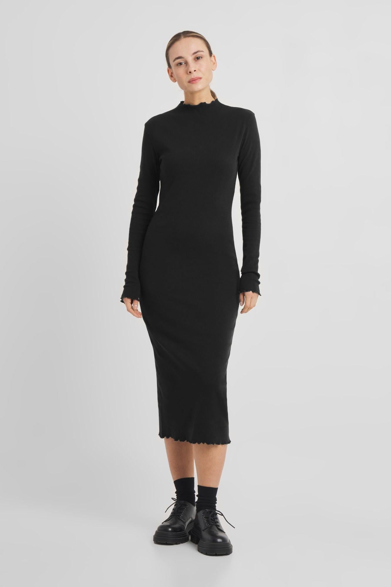 A woman modeling a black long-sleeve mock neck midi dress with a subtle textured knit and frayed hem, paired with black socks and black leather shoes, against a light gray studio background.