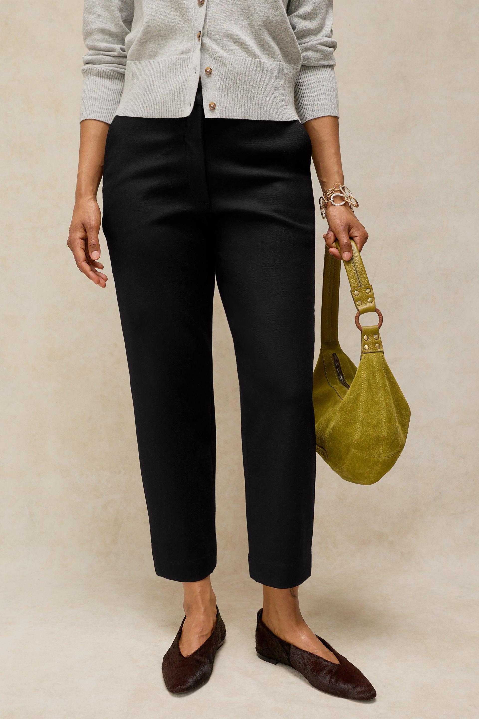 A model wearing black cropped trousers, a light gray cardigan, and dark brown pointed flats, holding a yellow-green suede shoulder bag against a textured beige background.
