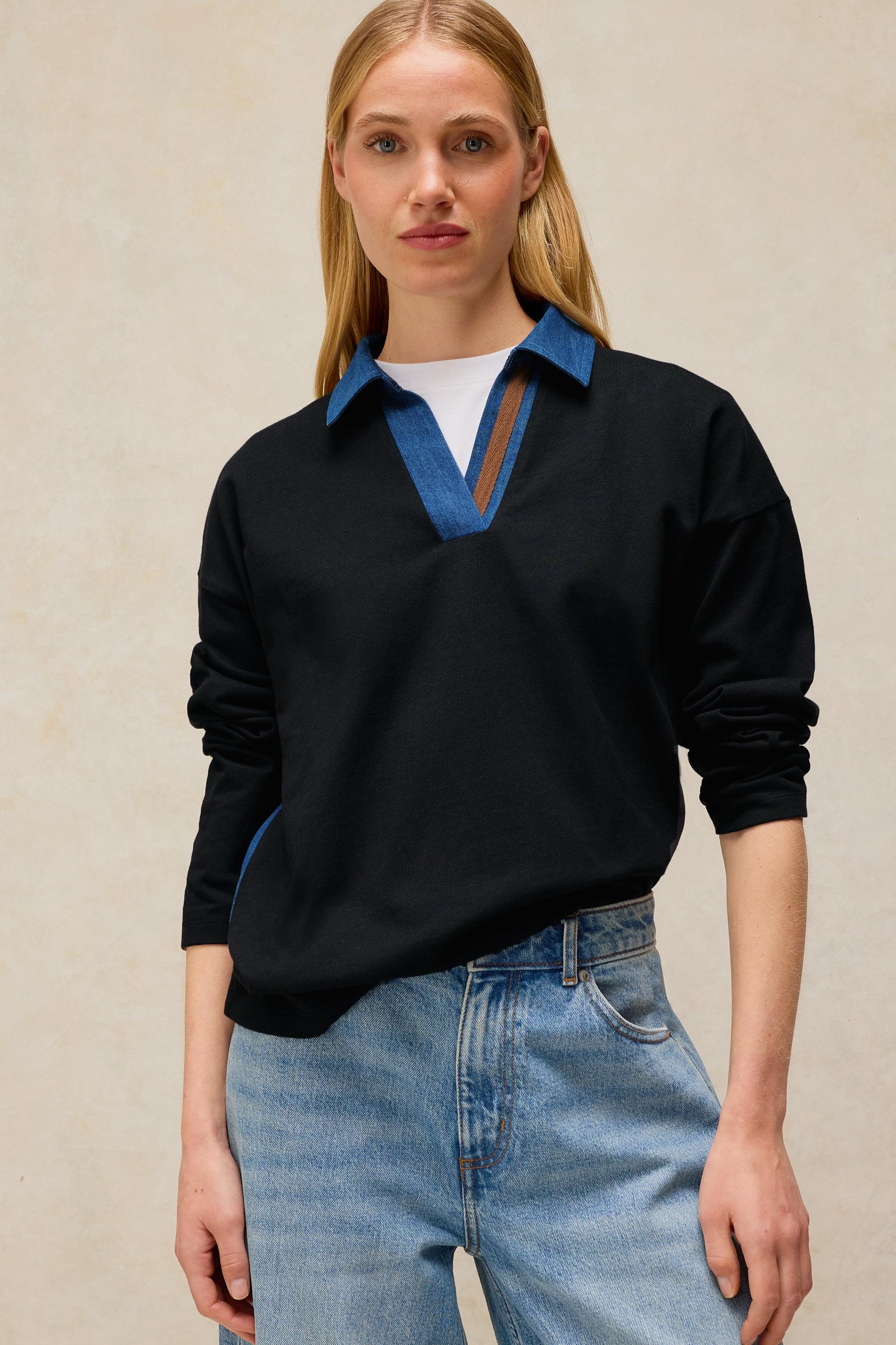 Woman wearing a black sweater with blue collar and jeans against a beige background