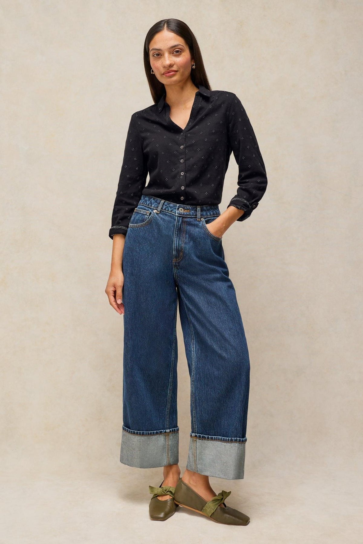 A woman posing in a black sparkle shirt and wide-leg cuffed denim jeans against a neutral studio background.