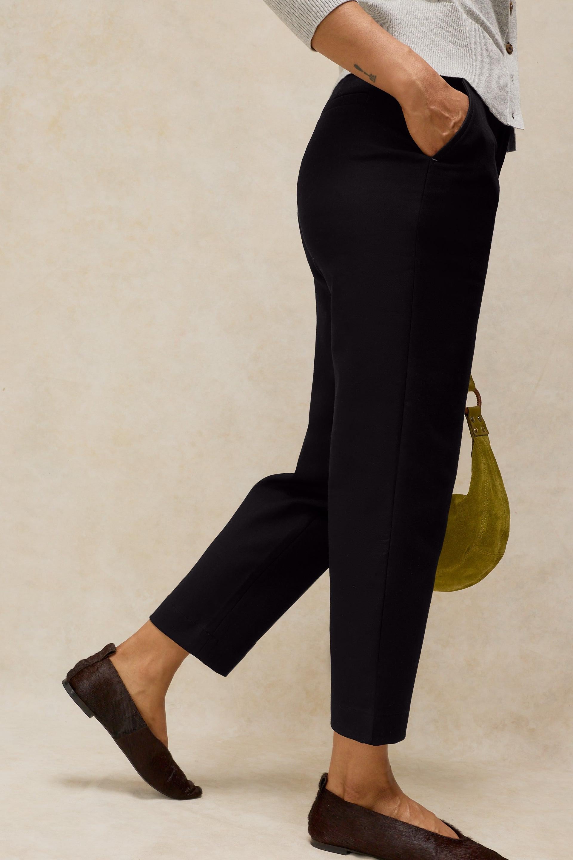 A person wearing black cropped trousers with side pockets, paired with a light gray top and dark brown flat shoes, standing against a textured beige background and holding a small olive green handbag.