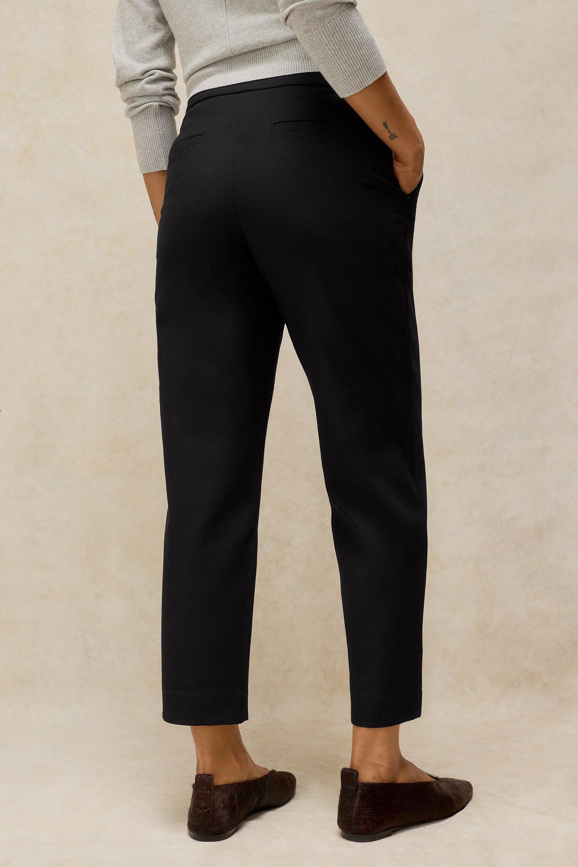 A person shown from the back wearing black cropped trousers, a light gray long-sleeve top, and dark brown flat shoes against a textured beige studio background.