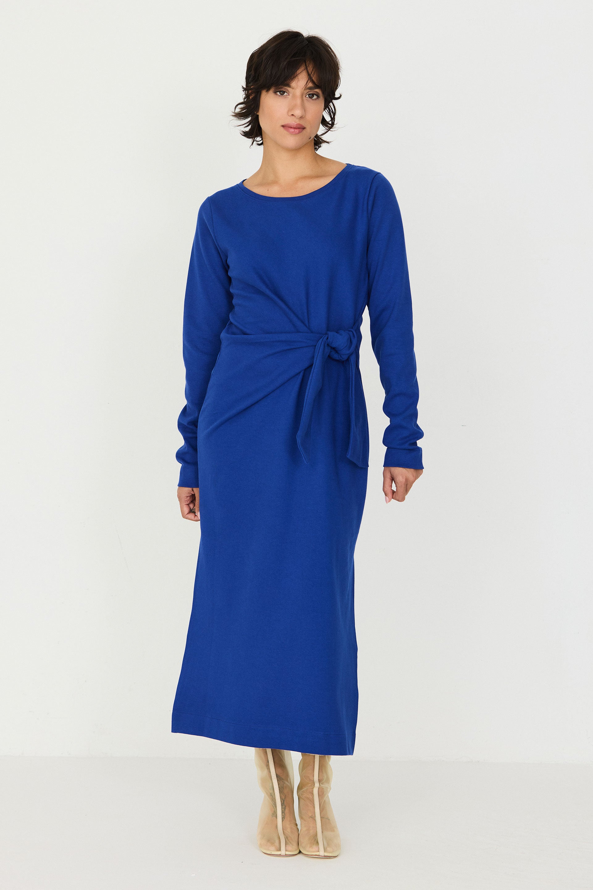 A woman modeling a long-sleeve midi dress in a vibrant royal blue color. The dress features a crew neckline, a wrap-style waist tied in a bow, and a draped side detail.