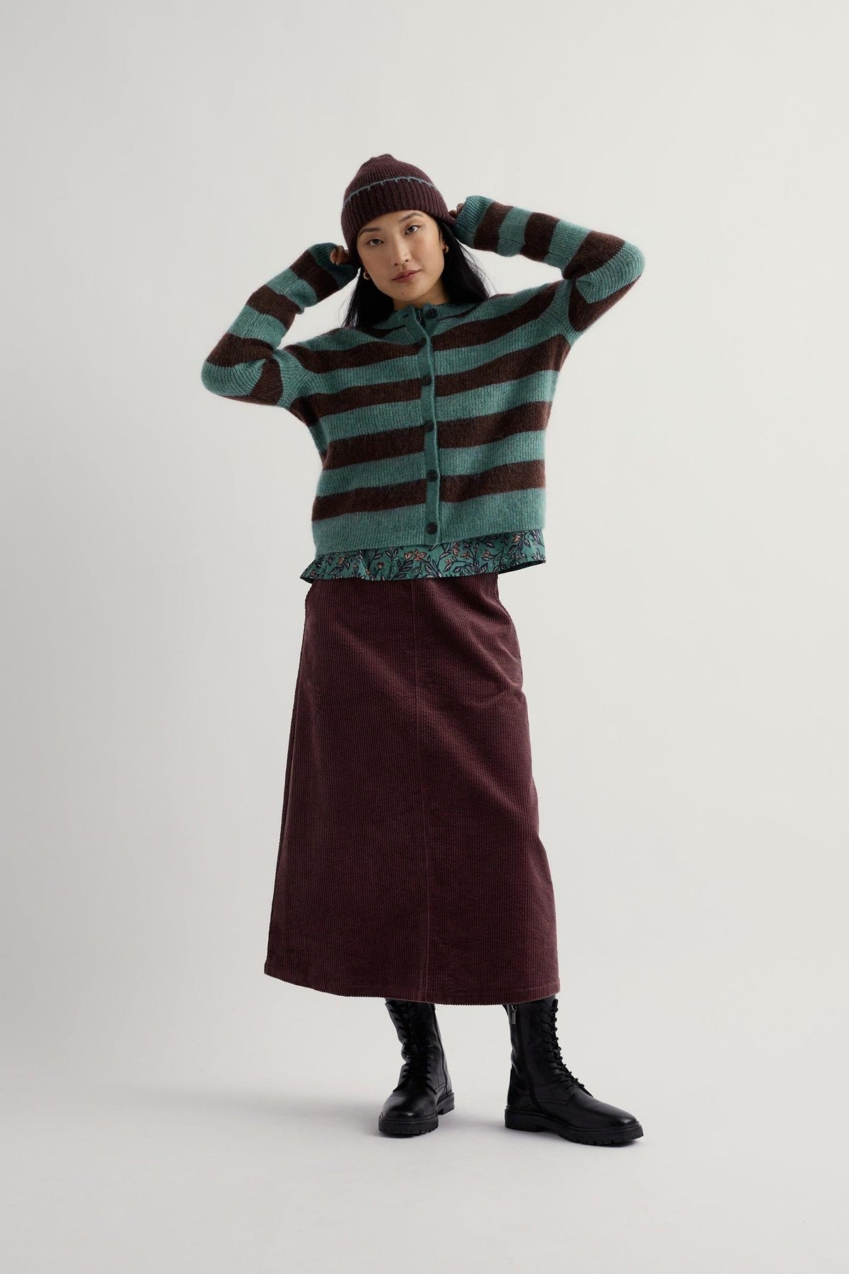 A woman posing in a blue and brown striped knit cardigan with a matching beanie and corduroy midi skirt.