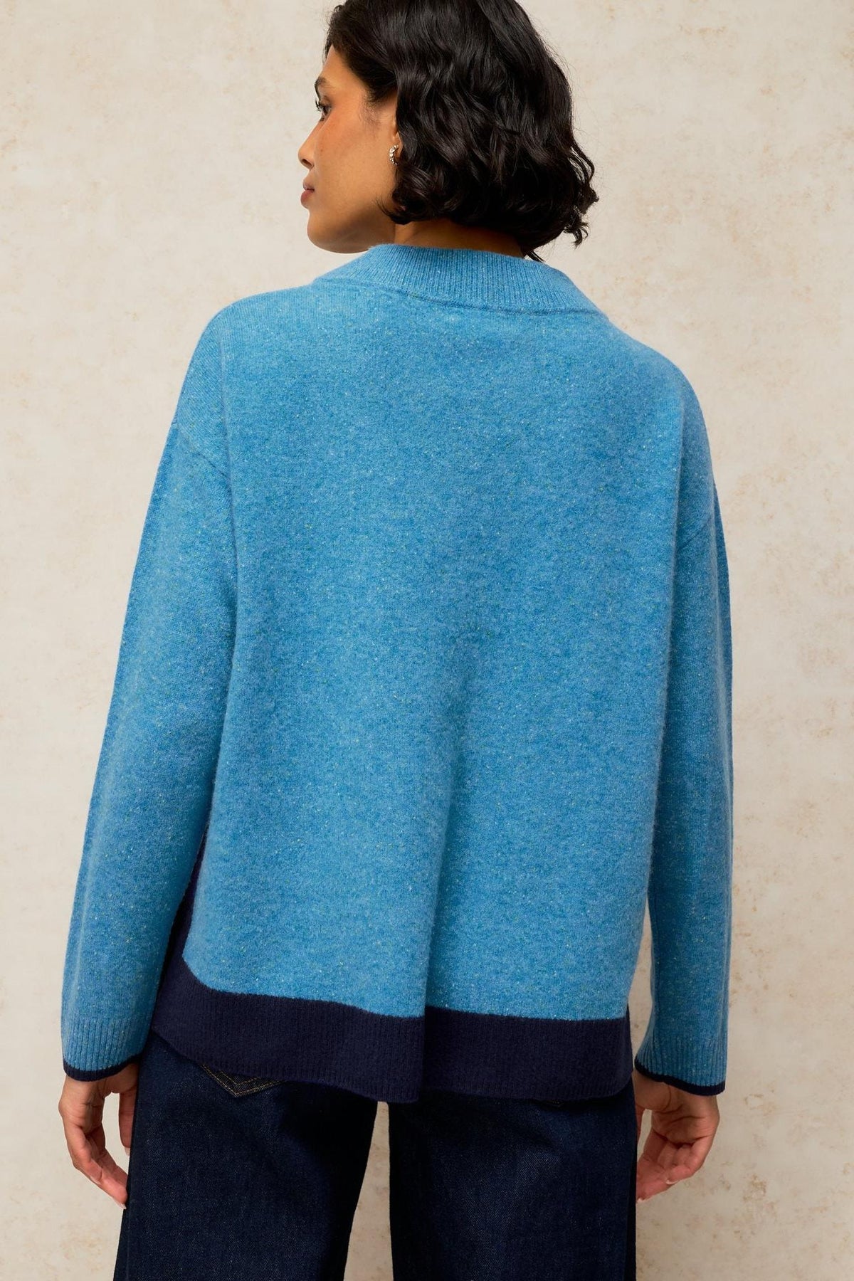 A woman seen from the back wearing a textured blue knit jumper with dark navy trim against a neutral studio background.
