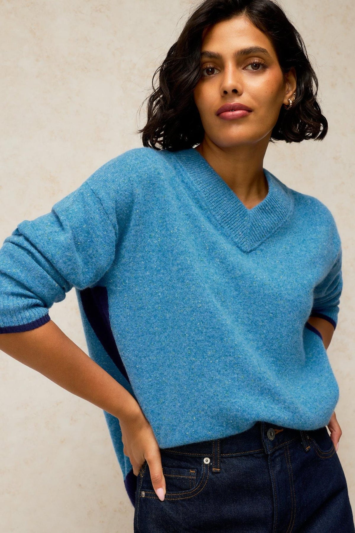 A woman posing in a vibrant blue v-neck knit sweater with dark denim jeans against a neutral background.