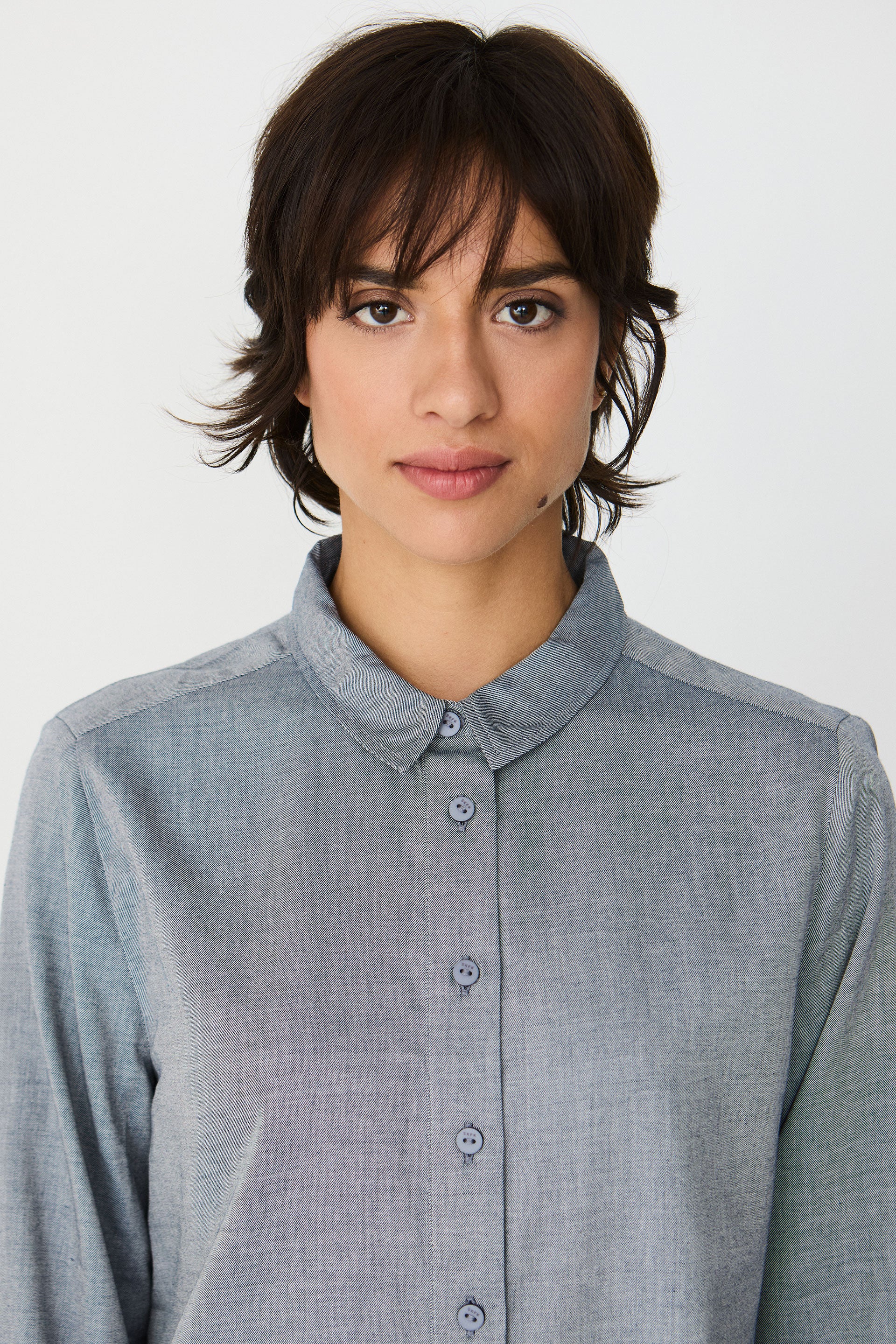 A woman with short brown hair wearing a buttoned blue melange shirt against a white background.