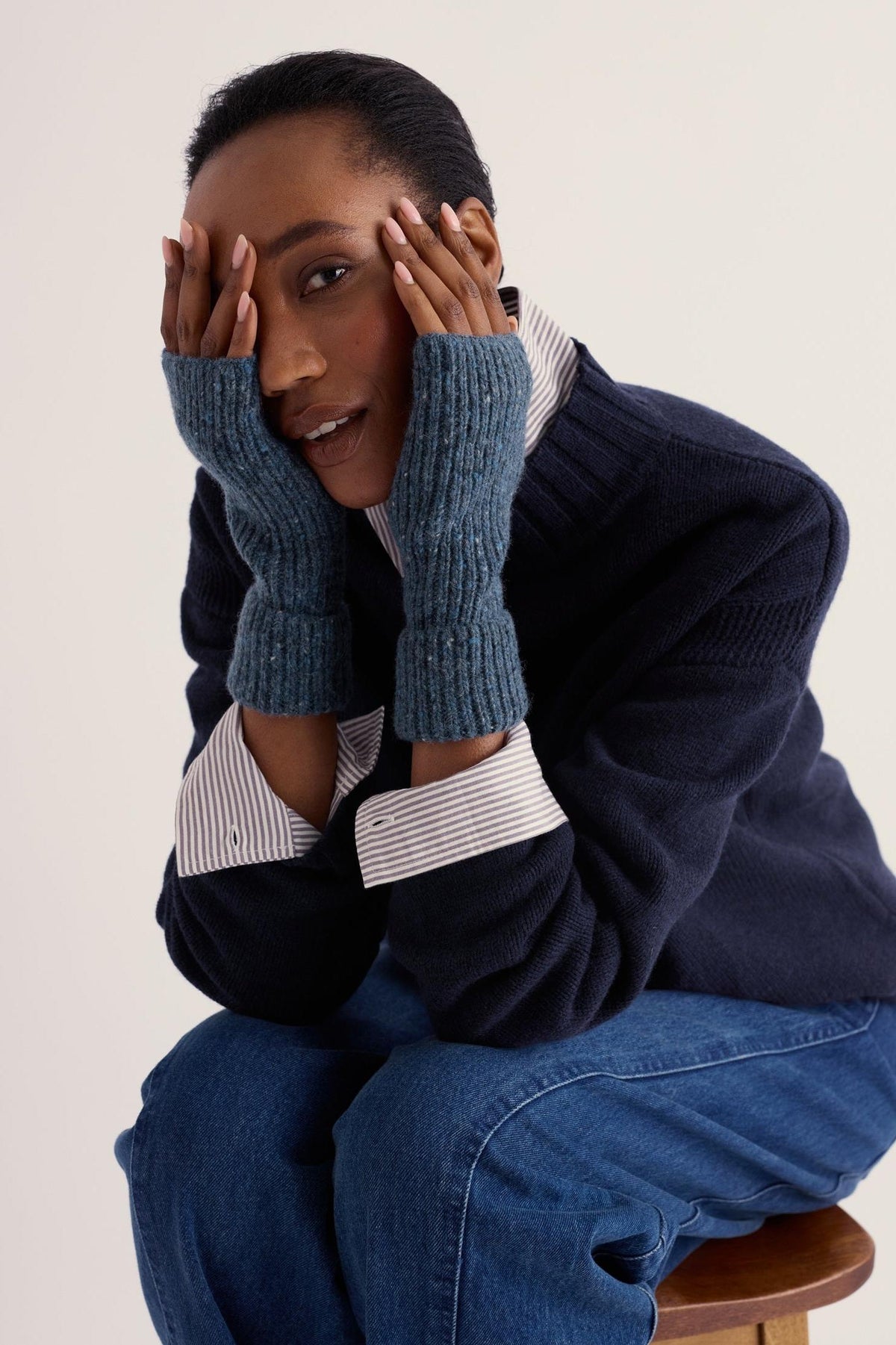 A woman wearing blue wool fingerless gloves covering her eyes against a neutral background.