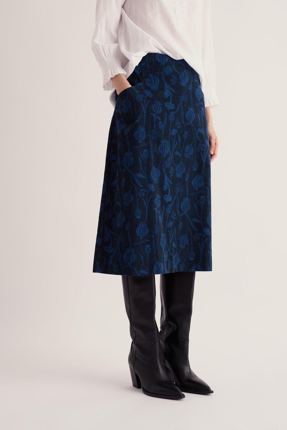 Woman wearing a blue needlecord long skirt with a floral pattern, paired with a white blouse and black boots.