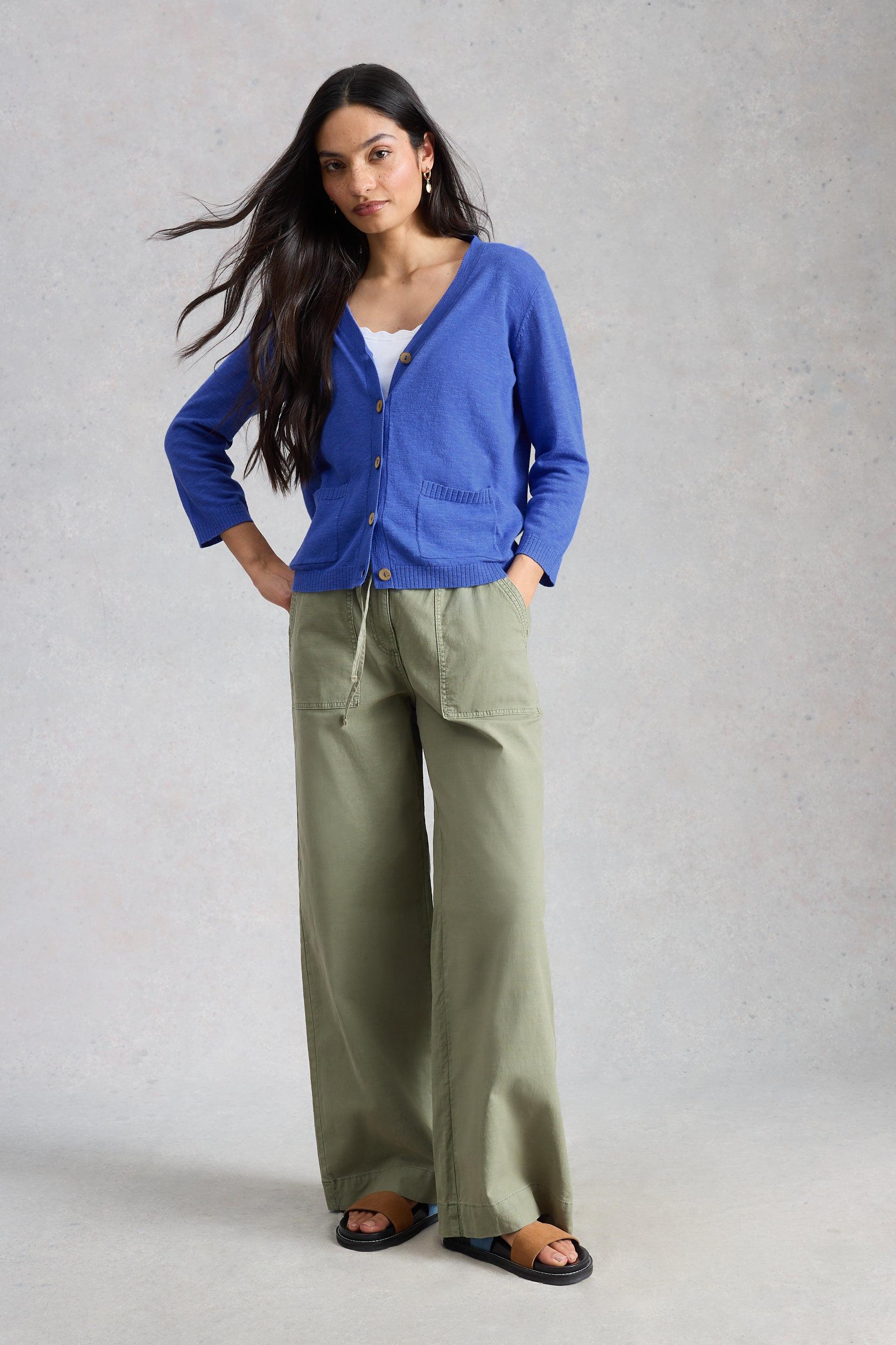 Woman posing in a blue organic cotton cardigan and green wide-leg trousers against a textured grey background.