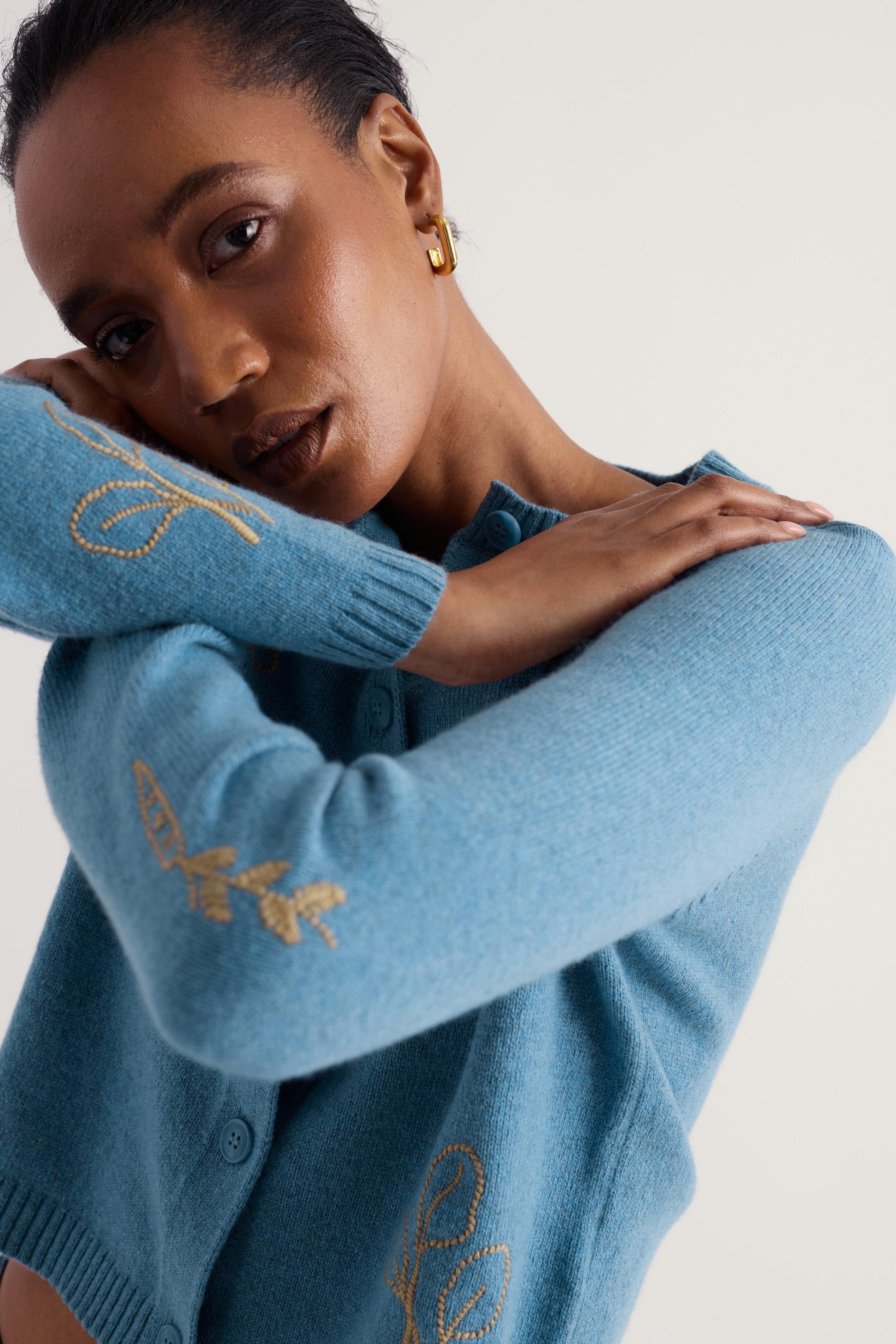 A close-up of a model wearing a light blue knit cardigan with gold embroidered leaf patterns on the sleeves and hem, posed against a plain light beige background.