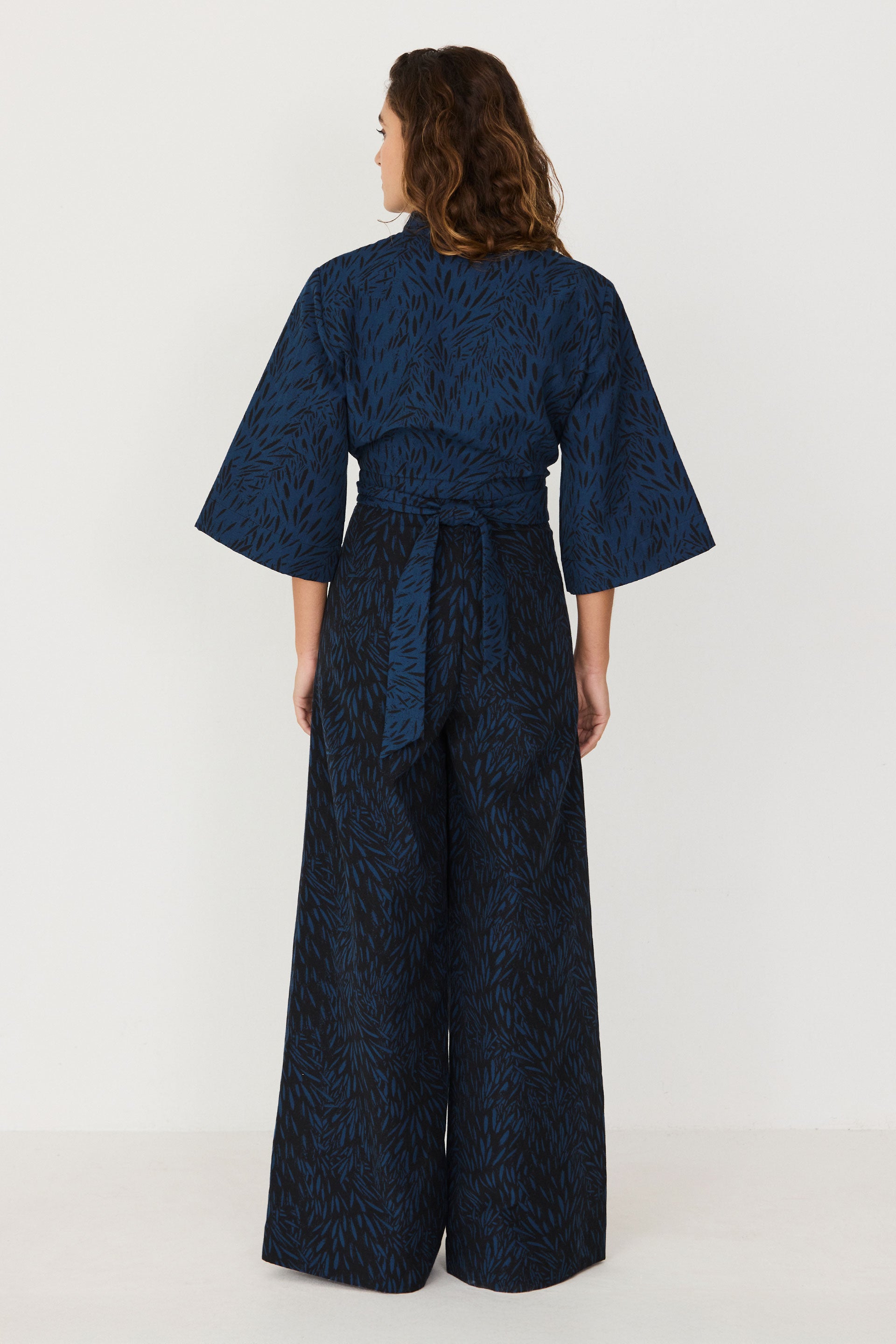 Back view of a woman wearing a blue organic cotton jumpsuit with a waist tie and wide-leg trousers