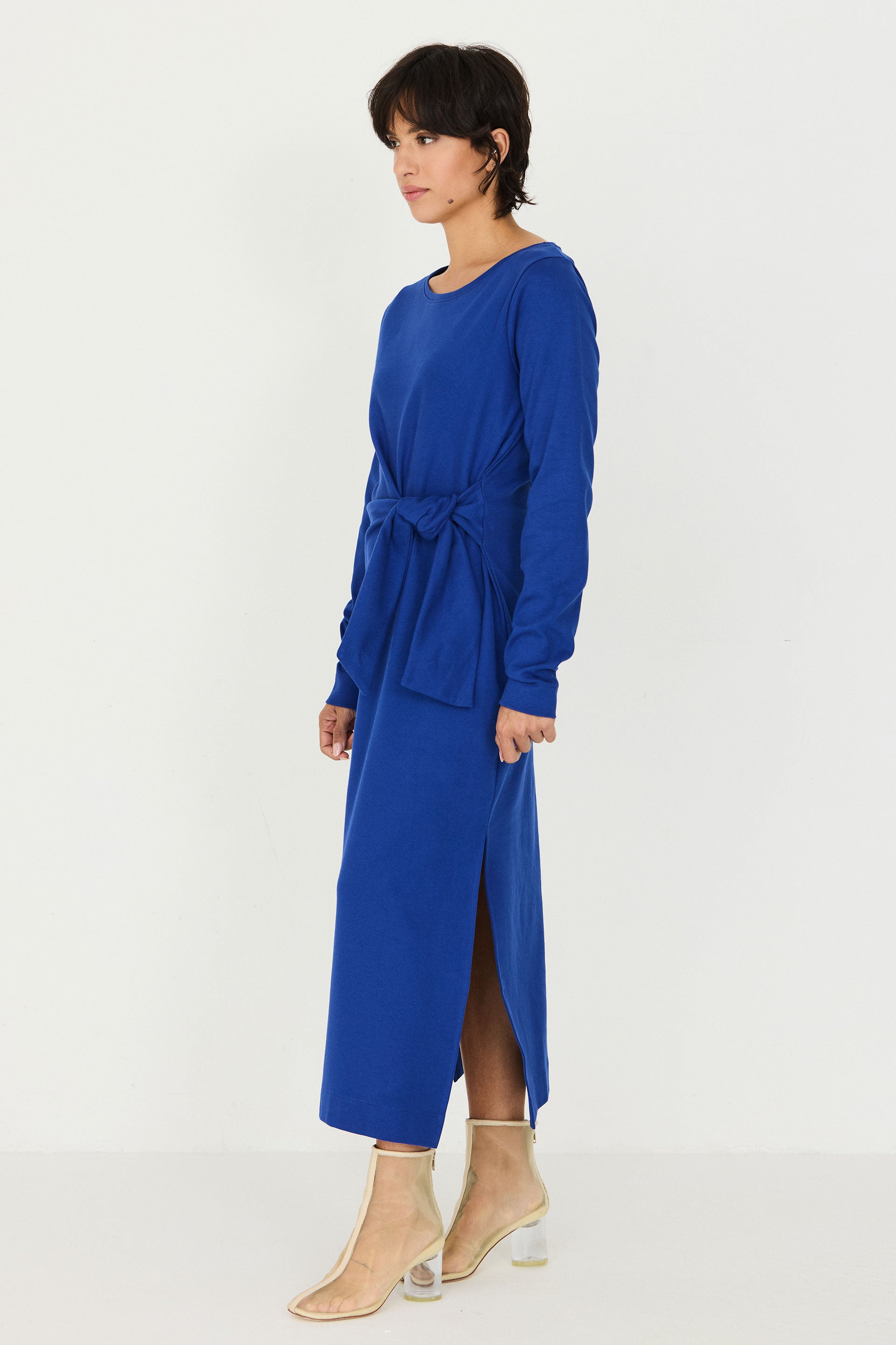 A woman modeling a long-sleeved midi dress in a vibrant royal blue colour, featuring a tie-waist belt and a front slit, paired with clear high-heeled sandals against a plain white studio background.