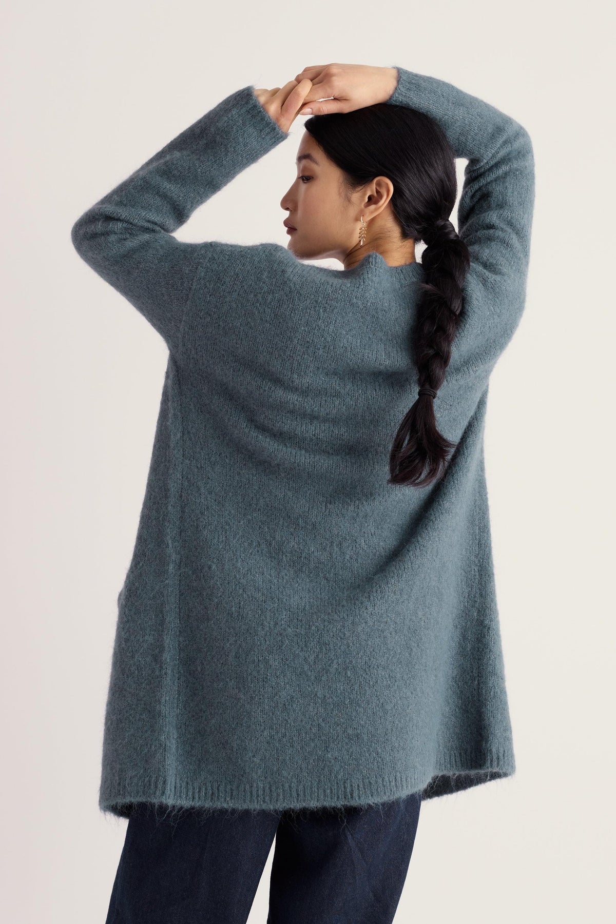 A woman wearing a long blue wool-blend cardigan with hands behind her head against a plain background.