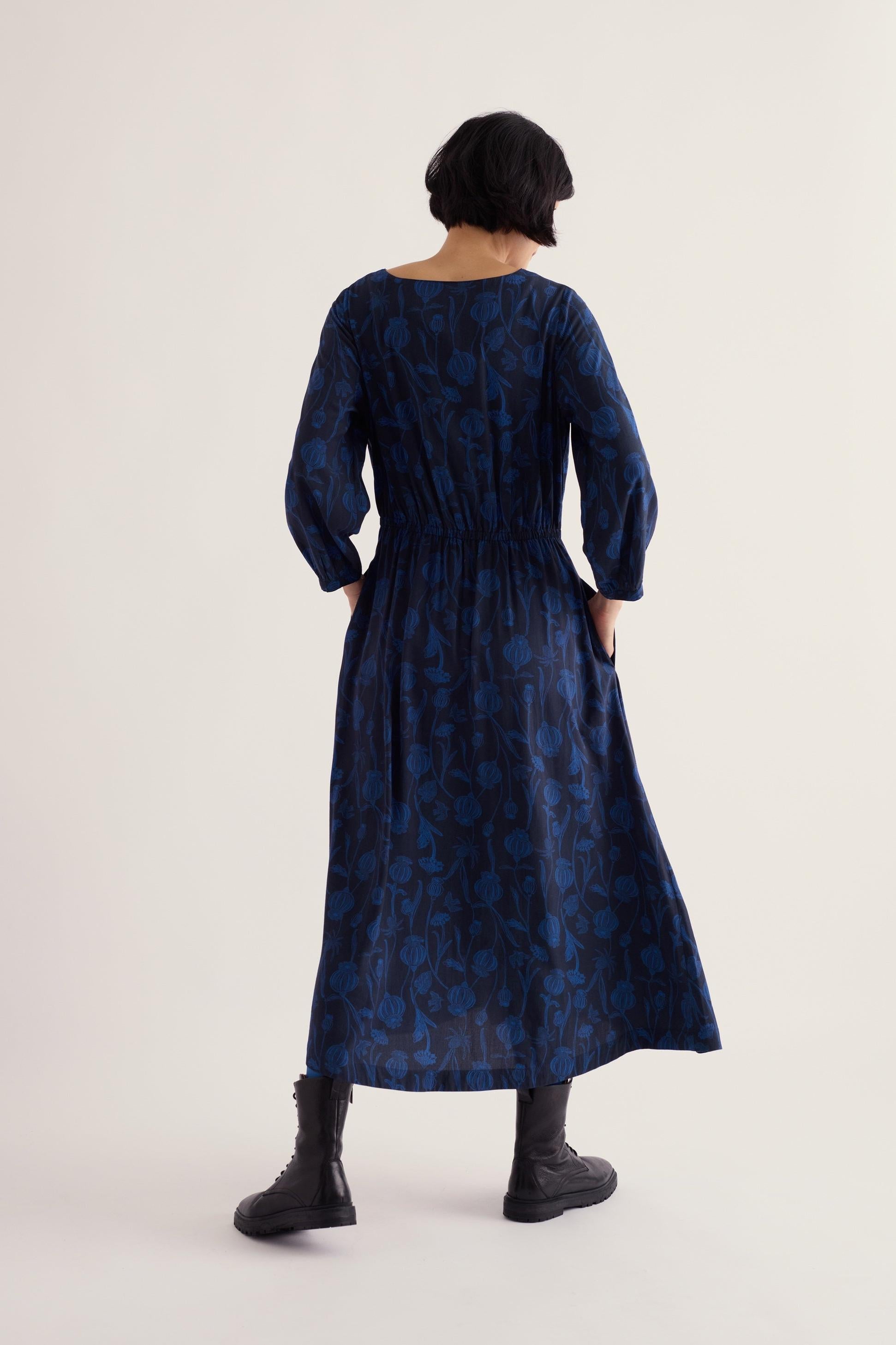 Woman seen from behind wearing a blue midi length Seasalt Cornwall dress with floral pattern and black boots.