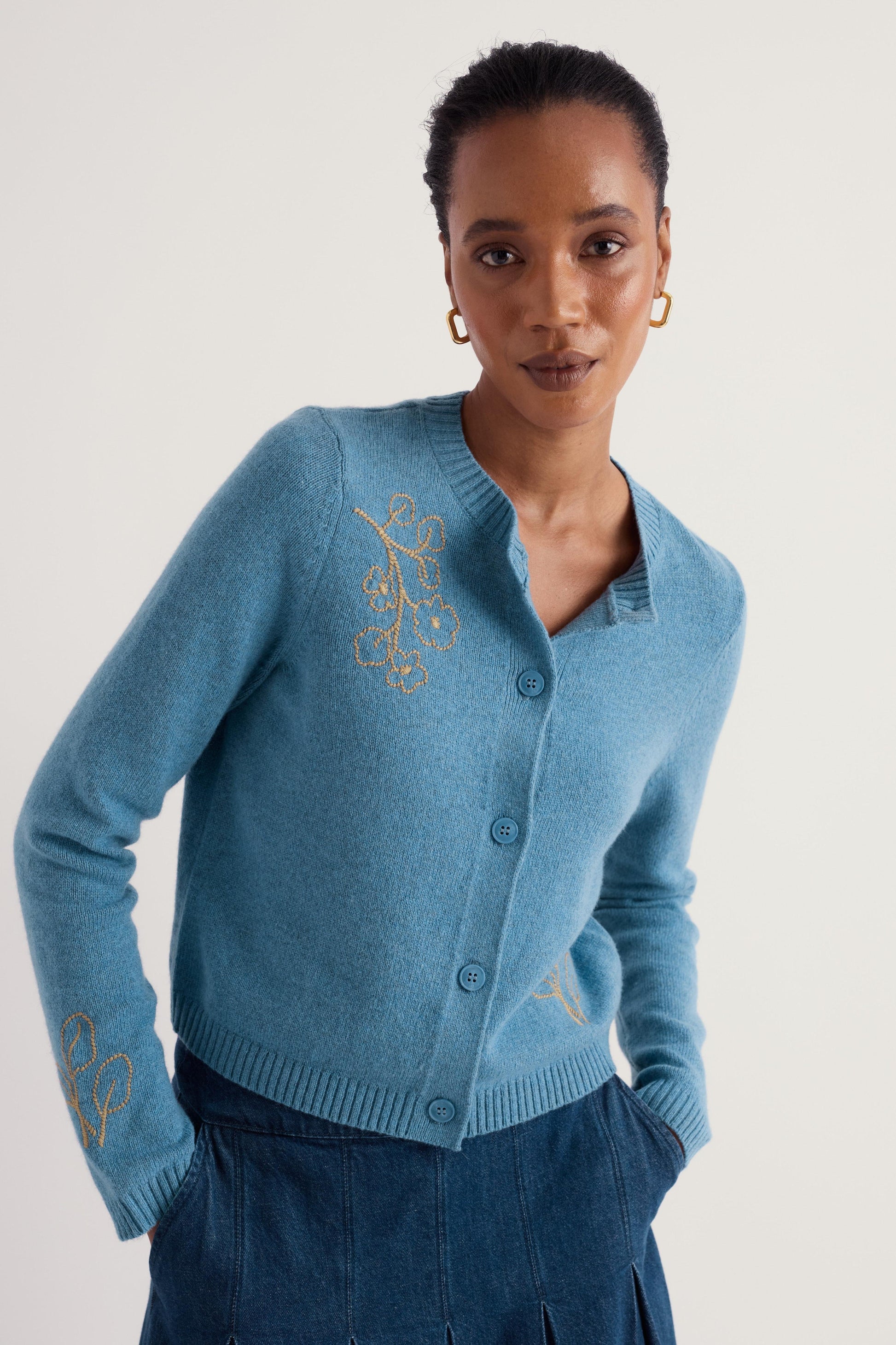 A woman wearing a light blue knit cardigan with gold embroidered details on the chest and sleeve, paired with dark blue trousers, against a plain light gray background.