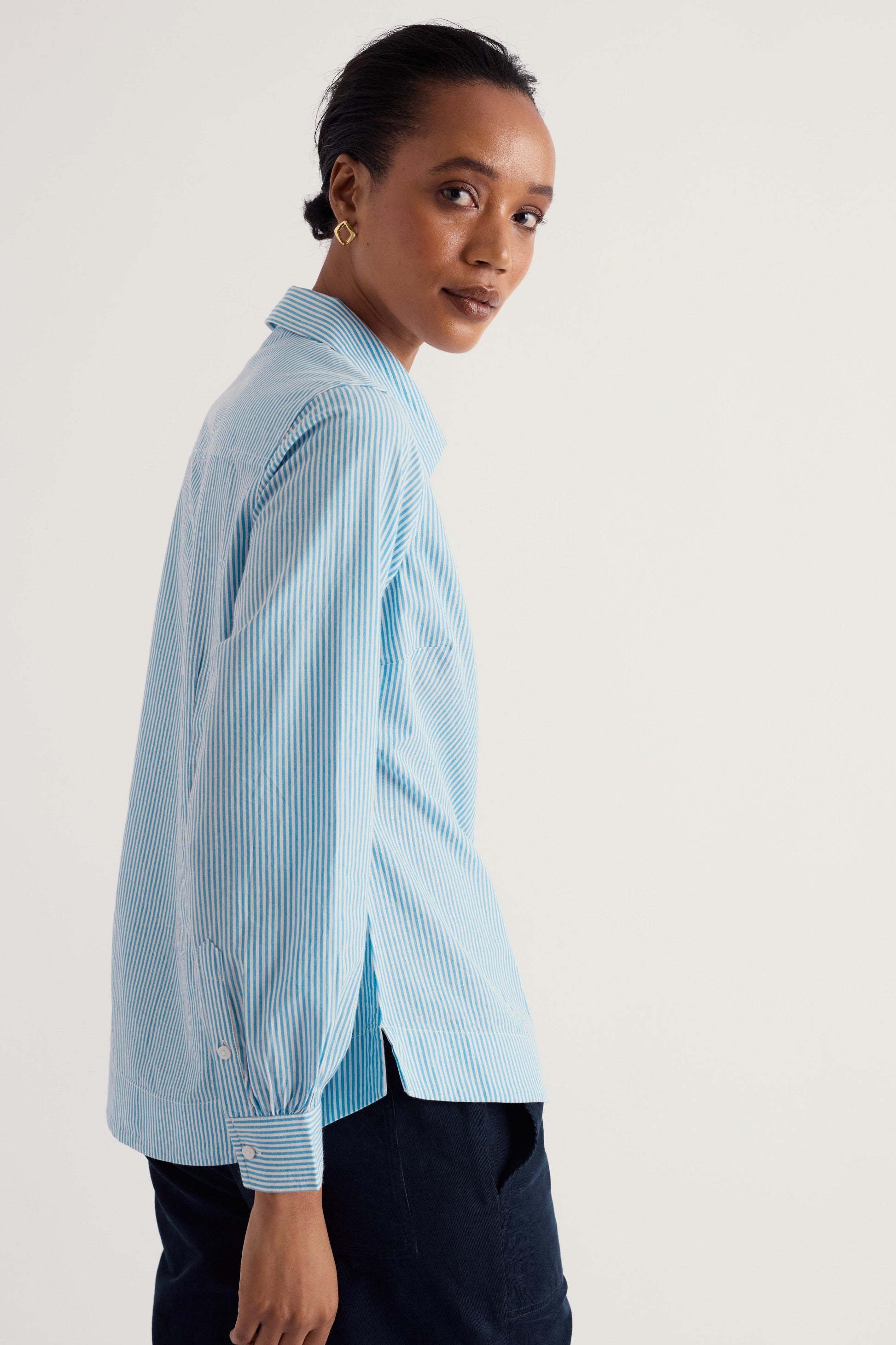 A woman wearing a light blue and white vertically striped button-down shirt, shown from a side-back angle against a plain off-white studio background.