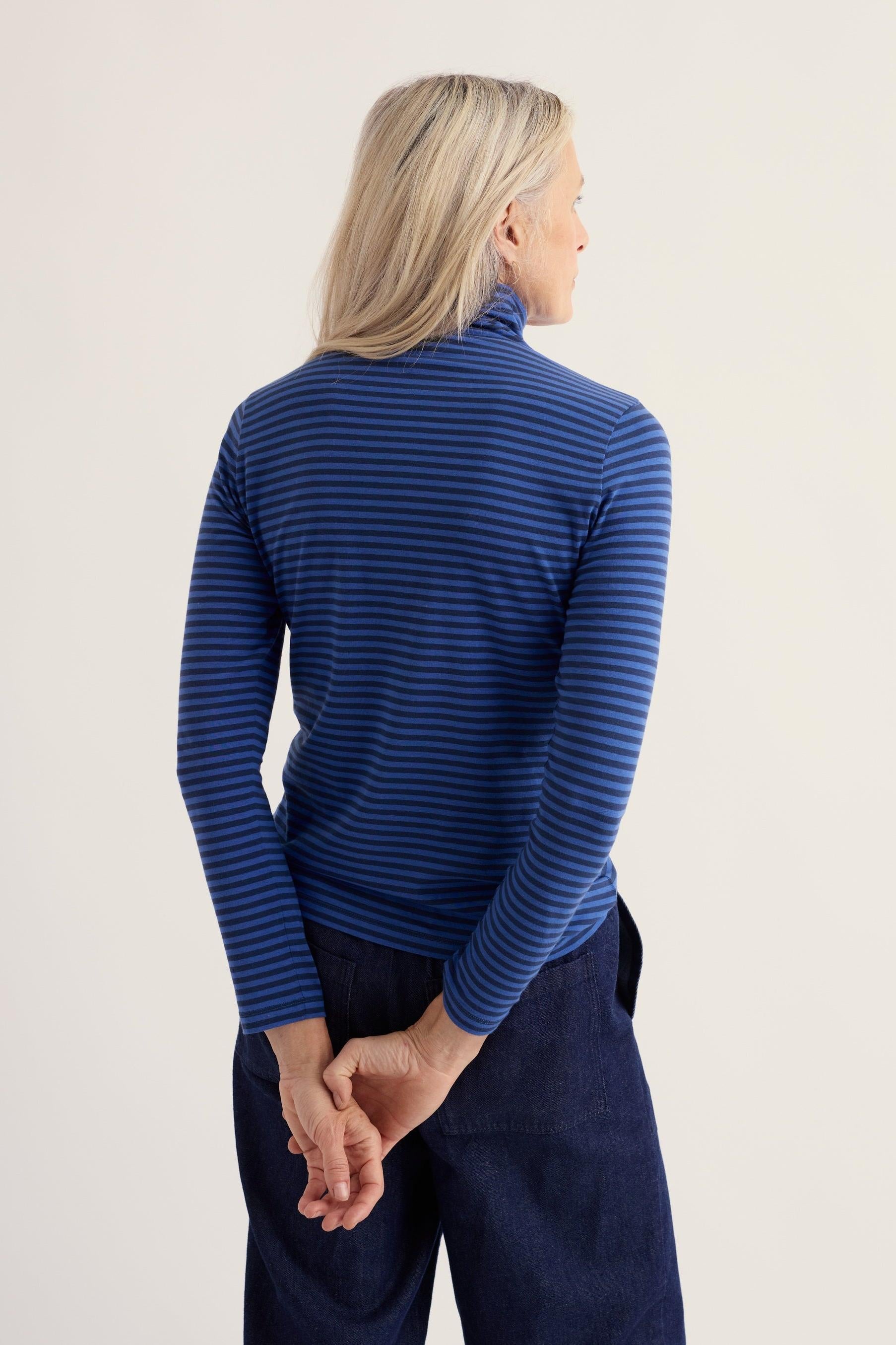 Woman seen from behind wearing a Seasalt Cornwall blue stripe organic cotton rollneck top with dark jeans.