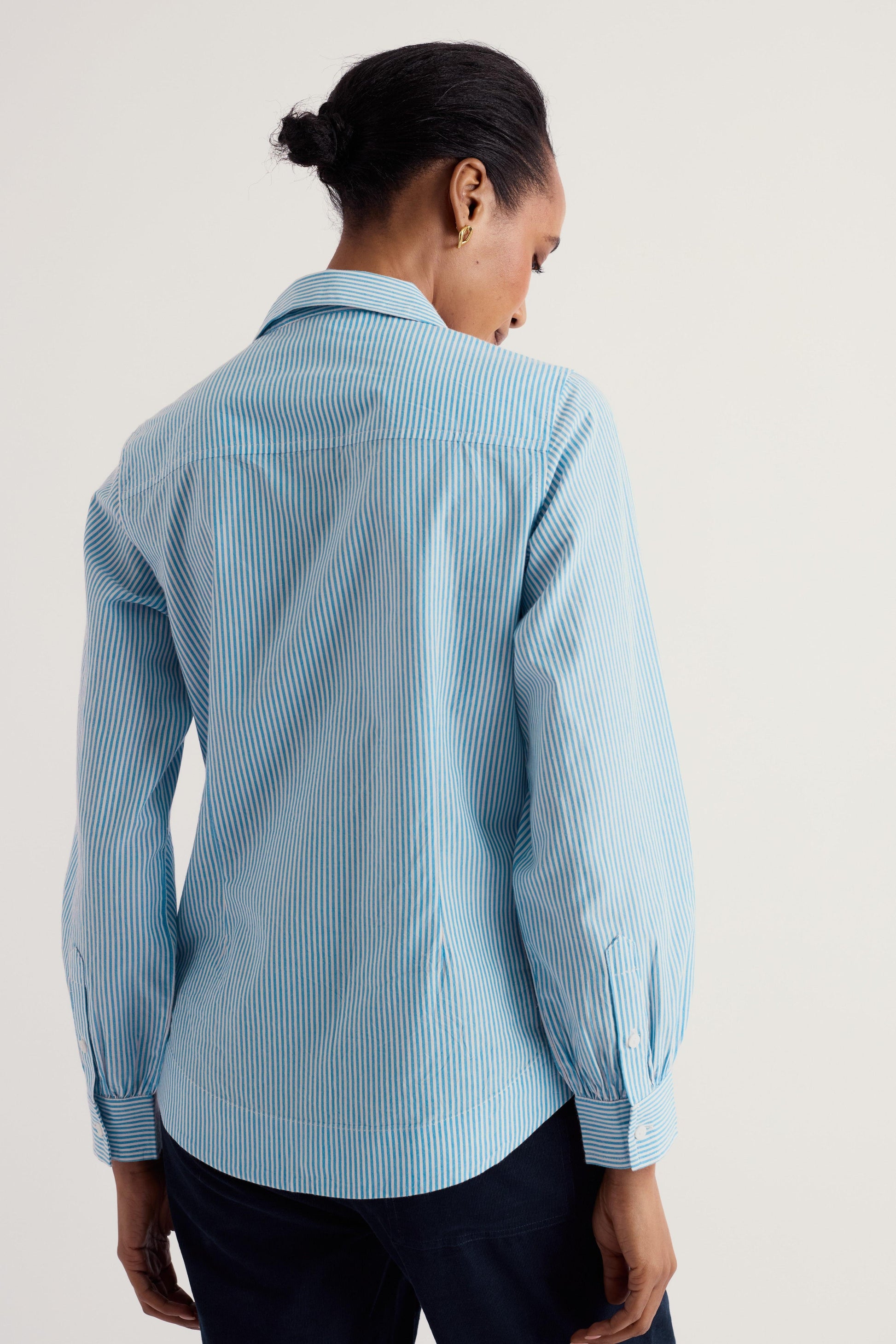 A woman shown from the back wearing a light blue and white vertically striped button-down shirt, paired with dark trousers, against a plain off-white studio background.