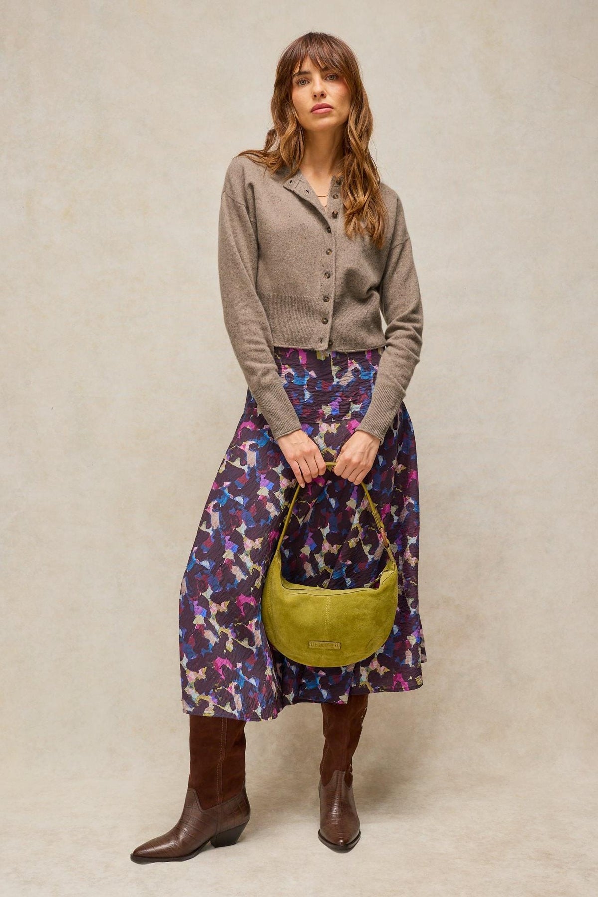 A woman posing in a taupe cashmere cardigan, a colourful printed midi skirt, and brown boots holding a lime green bag.